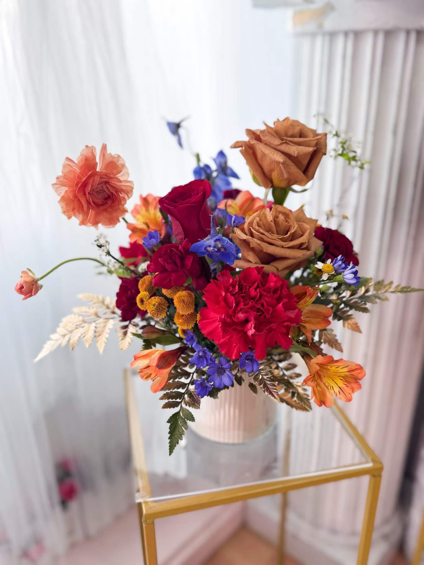 [FRESH FLOWER] Amber Velvet Garden - A rich and artistic floral arrangement featuring luxurious caramel roses, vibrant crimson blooms, soft peach ranunculus, delicate blue delphinium, and seasonal accent flowers in warm sunset tones. Designed in a modern ribbed ceramic vase, this arrangement combines earthy elegance with a whimsical garden-inspired style. The unique caramel roses add a sophisticated neutral warmth that beautifully contrasts with the vivid reds and soft blue accents, creating a design that feels both modern and romantic. Perfect for birthdays, celebrations, thank you gifts, home décor, or anyone who appreciates distinctive floral artistry. Each arrangement is handcrafted with premium seasonal flowers. Due to seasonal and market availability, certain flowers may be substituted with blooms of equal or greater value while maintaining the overall color palette and overall style of the design.