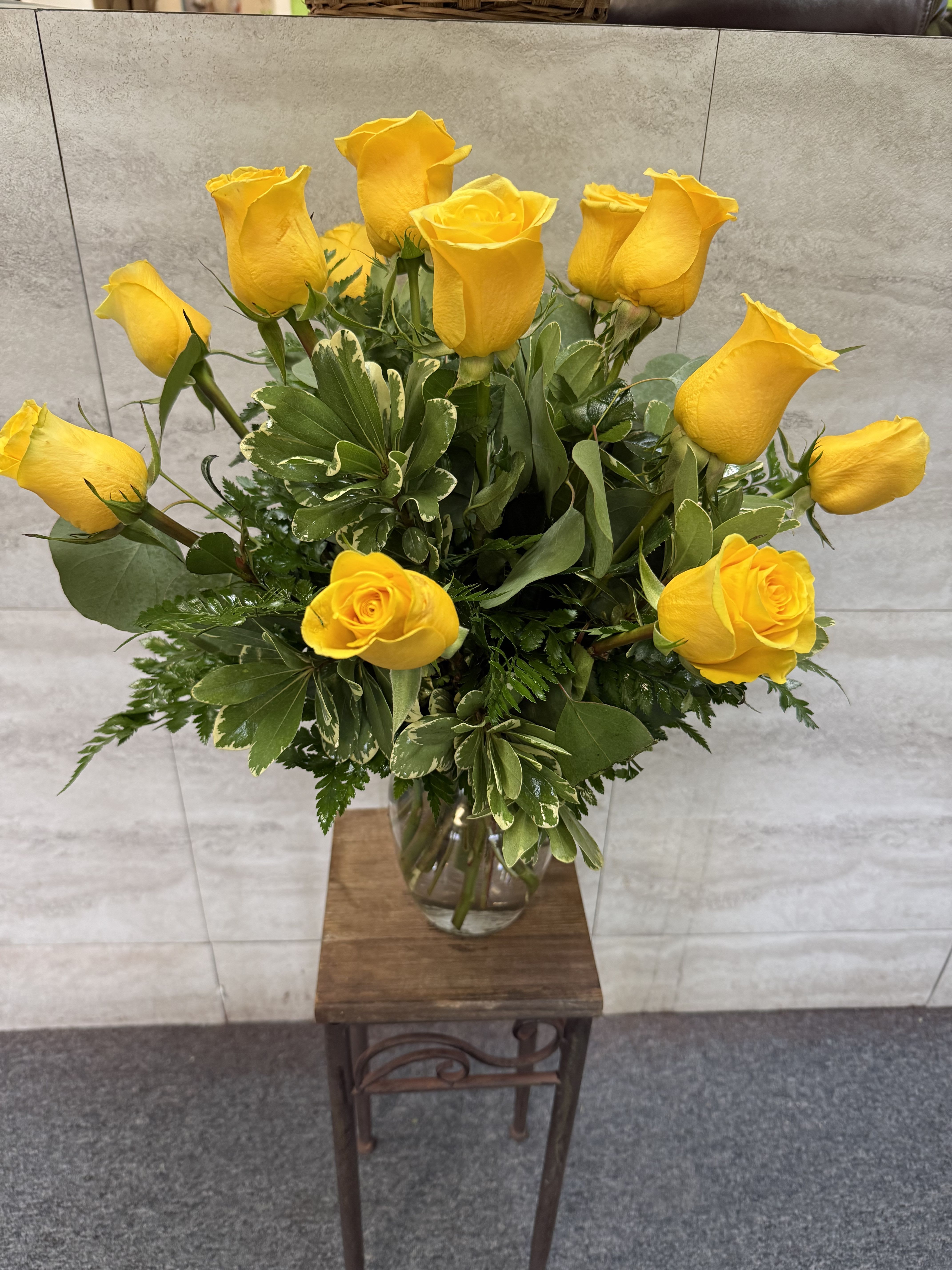 Tall arrangement of yellow roses in a clear glass vase on a wooden stand.