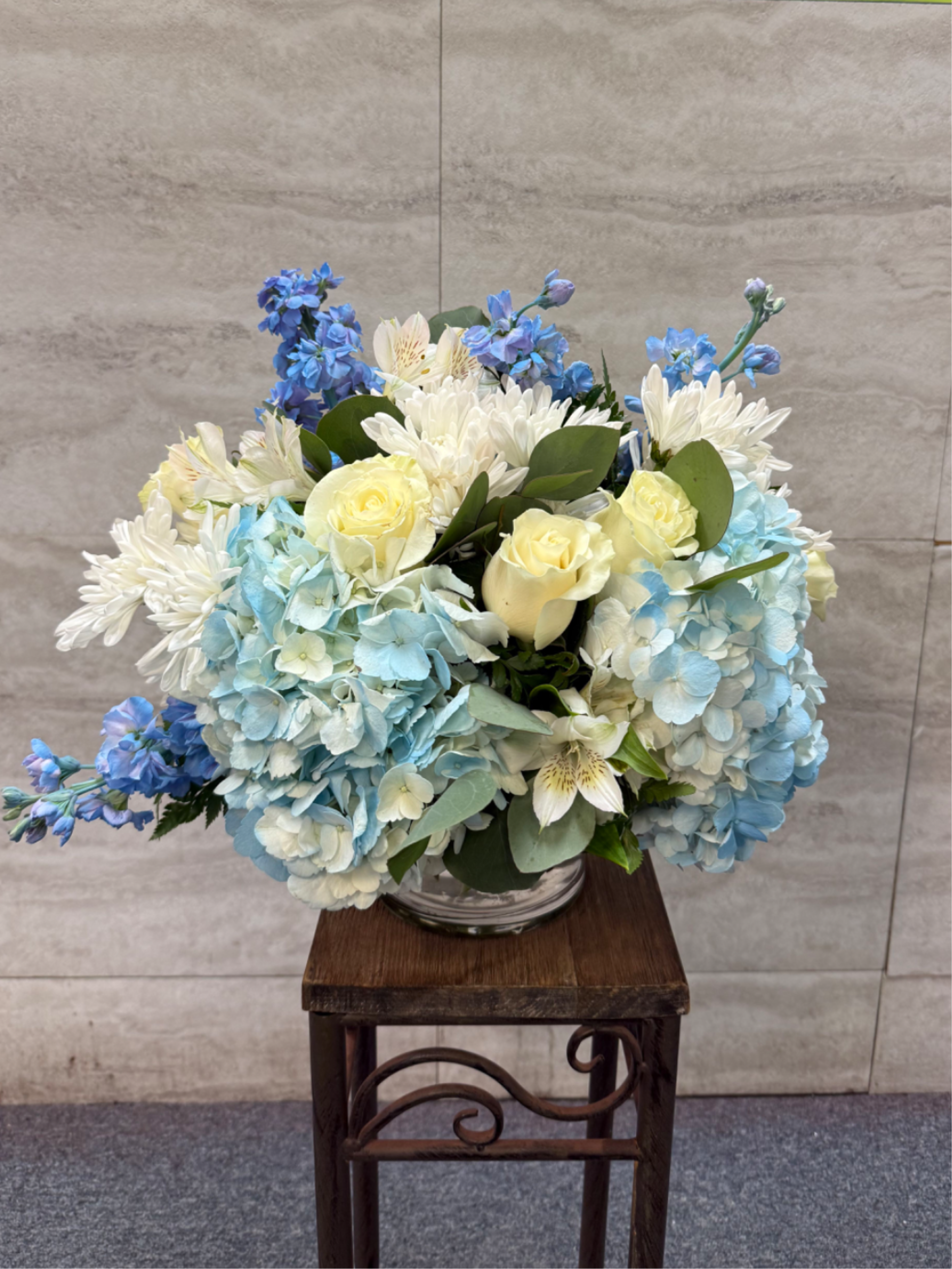 Blue Gem  - (ONE-SIDED) is a calm, elegant arrangement that feels fresh and effortlessly refined   Soft blue hydrangea form a lush foundation, accented by creamy white roses and crisp white chrysanthemums for a timeless, graceful look. Delicate blue delphinium and pale alstroemeria add light, airy movement, while rich greenery ties everything together with natural balance.  Cool, soothing, and beautifully polished, *Blue Gem* is perfect for expressing serenity, appreciation, and quiet sophistication—an arrangement that shines with gentle beauty rather than bold drama. 