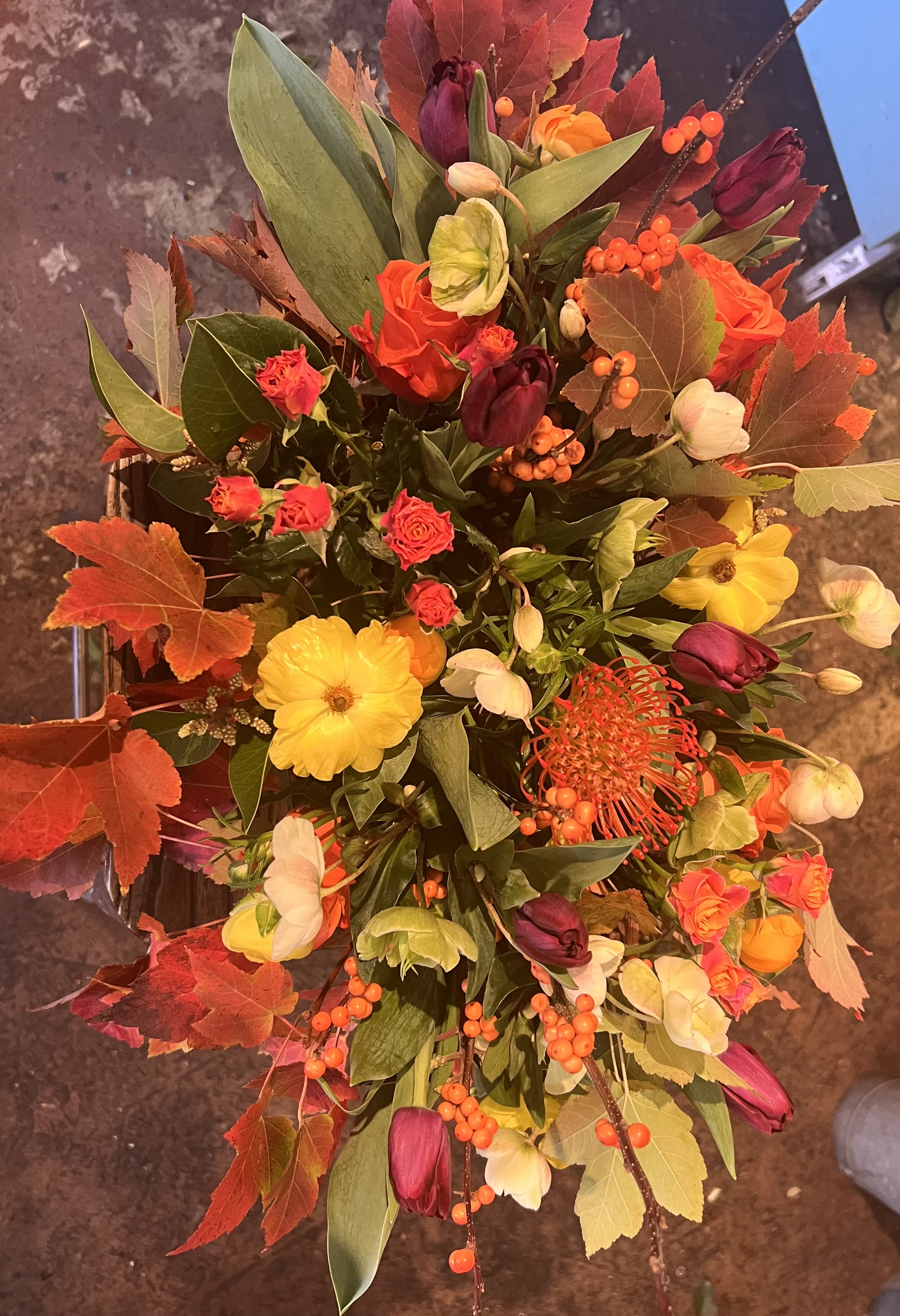 Autumn Ember - Warm- lush A vibrant seasonal arrangement featuring rich burgundy tulips, glowing orange roses, golden blooms, and playful berries. Layered with fiery autumn foliage and lush greenery, Autumn Ember brings warmth, texture, and true fall richness to any setting.