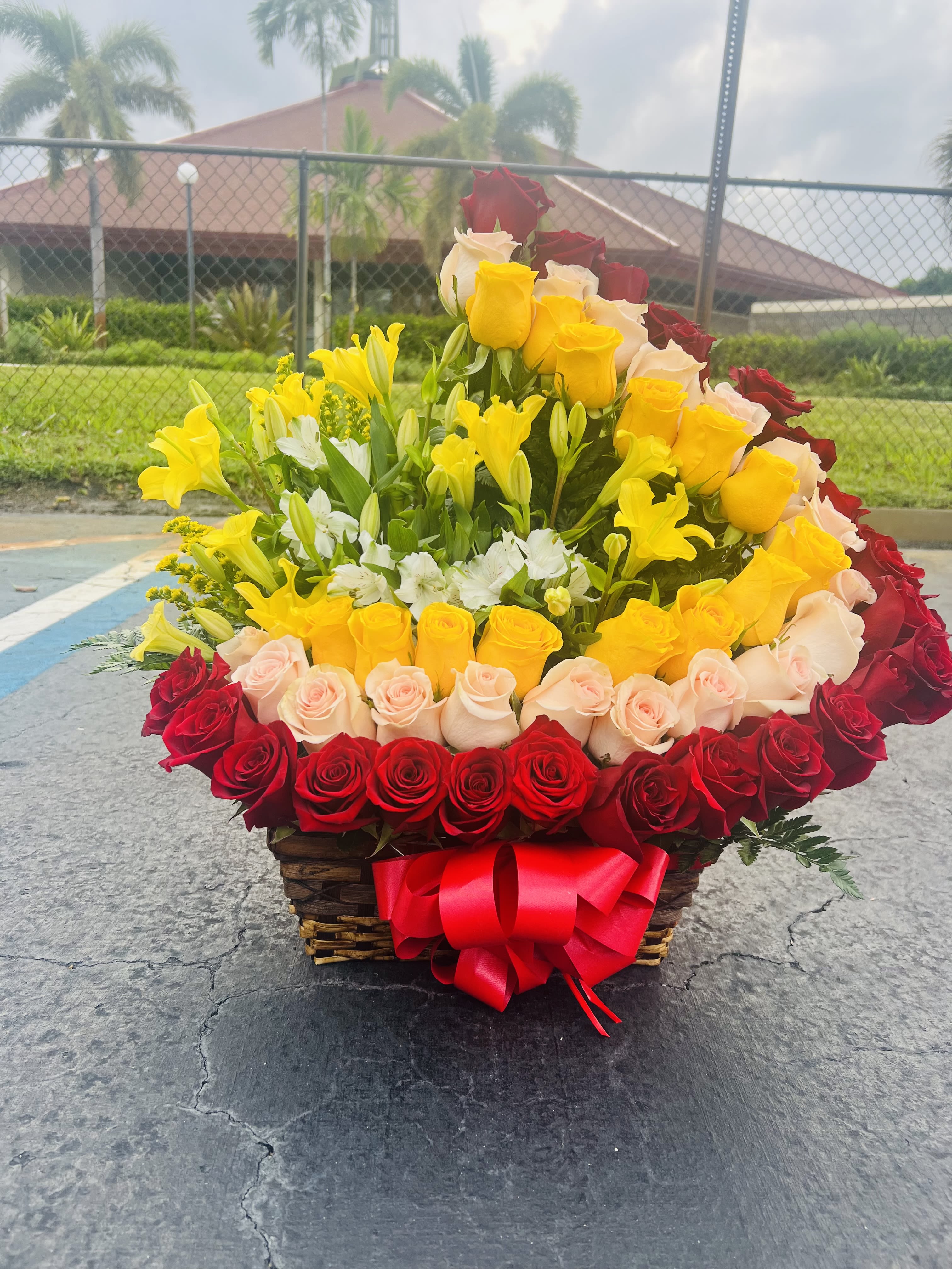 Basket arrangement of red, yellow, and blush roses with lilies and a red bow