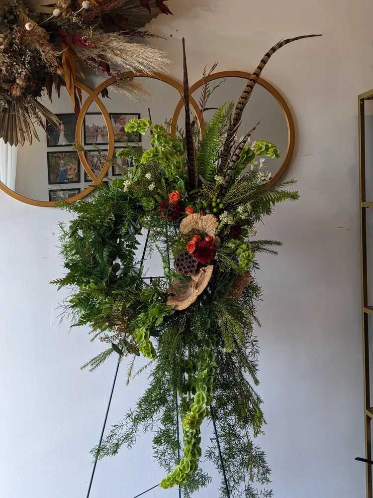 Nature’s Embrace Standing Spray - Created for those who felt most at home in nature, this standing spray is a heartfelt tribute inspired by the quiet beauty of the outdoors. Designed with layered greenery, organic textures, and seasonal blooms in rich, earthy tones, this arrangement captures the natural movement and balance found in the wild. Thoughtful details and flowing elements create a grounded yet graceful presence, offering comfort while honoring a deep connection to the natural world.  Ideal for funeral services, memorials, or celebration-of-life ceremonies. Flower varieties and natural elements may vary based on seasonal availability, while maintaining the overall look, tone, and intention of the design.