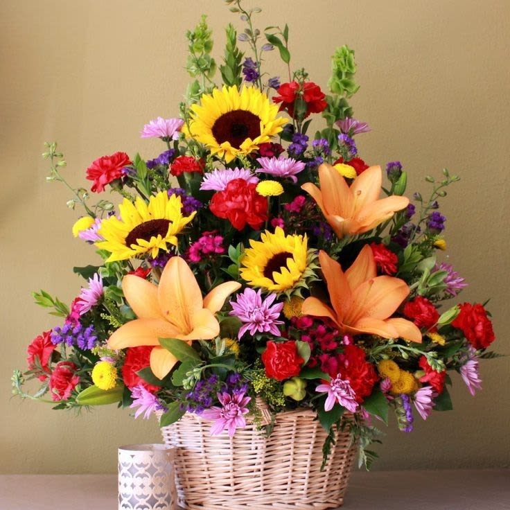 Colorful Garden Celebration - A lively, mixed-flower basket that feels like a sunny garden in miniature-perfect as a birthday bouquet, get well arrangement, or a "thinking of you" gift. This flower arrangement pairs bright yellow sunflowers and blue delphinium with a cheerful mix of roses, carnations, lilies, alstroemeria, chrysanthemums and daisies to create a bold, celebratory look that still reads warm and homey. The fan-shaped composition and tall Bells of Ireland add pleasing height so the recipient feels genuinely noticed.  Arranged in a woven wicker basket with a small decorative votive holder for scale, the piece balances bold color-yellows, oranges, reds, pinks and purples-with calming white clusters to keep the design fresh and versatile. Textures range from the soft rose and carnation petals to the spiky delphinium and structured sunflower heads, making the bouquet feel abundant without being fussy. Sized for tabletop display, it's an easy way to brighten a hospital room, bedside, office desk, or entry table.  Choose this arrangement when you want someone to feel celebrated, cheered, or comforted; it reads festive enough for congratulations yet gentle enough for get-well or sympathy. Arrives ready to display in its wicker basket-swap the votive for a candle or keep it as shown-and refresh water daily to keep blooms at their best.