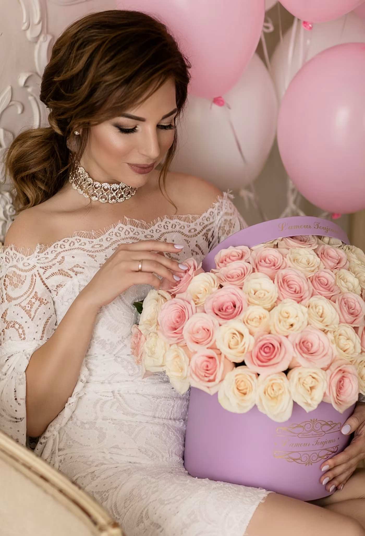 Woman in a white lace dress holding a box of pink and cream roses with balloons behind her