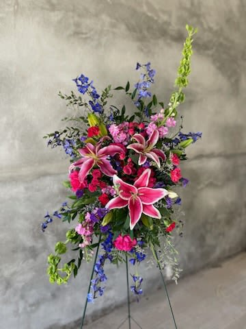Radiant Rememberance Spray - A bold and beautiful tribute, Radiant Remembrance Spray is designed to celebrate a life full of color, love, and cherished memories. Featuring striking pink lilies, vibrant carnations, rich blue delphinium, and soft complementary blooms, this standing spray brings both elegance and heartfelt warmth to any service.  With its natural garden-style movement and uplifting color palette, this piece offers a meaningful expression of sympathy, remembrance, and honor.  > Colors and exact flowers may vary slightly based on seasonal availability > Designed fresh daily using the best blooms available > Each piece is uniquely handcrafted while maintaining the overall style