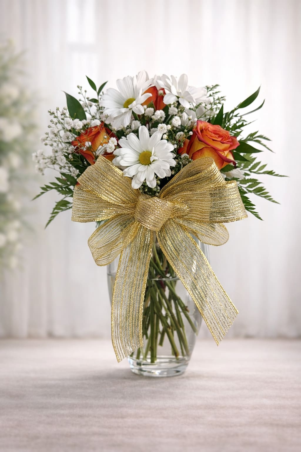 Bouquet of orange roses and white daisies in a clear glass vase with a gold bow