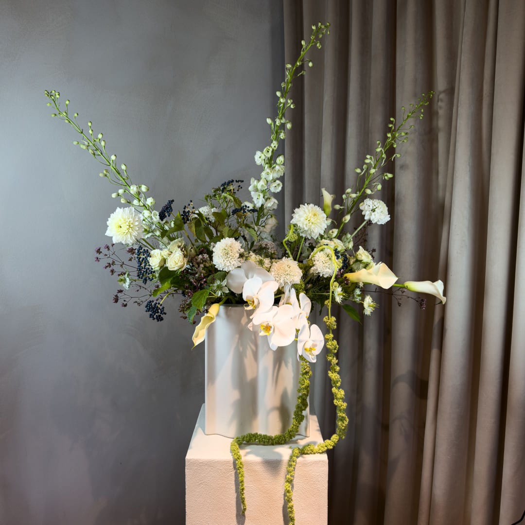 Tall floral arrangement with white orchids, calla lilies, and pale blooms in a white vase