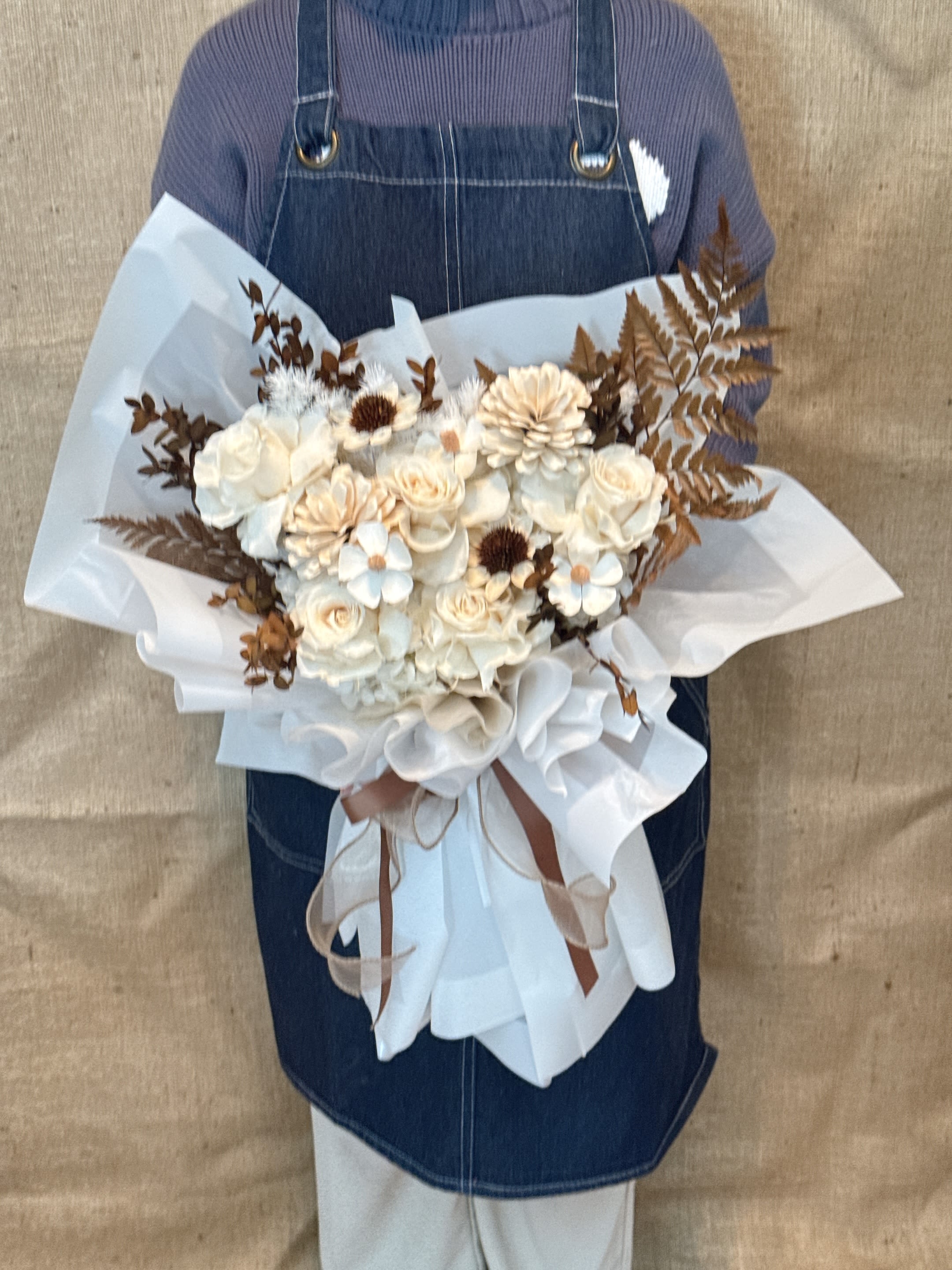 Handheld bouquet of cream roses and white blooms with brown foliage, wrapped in white paper.