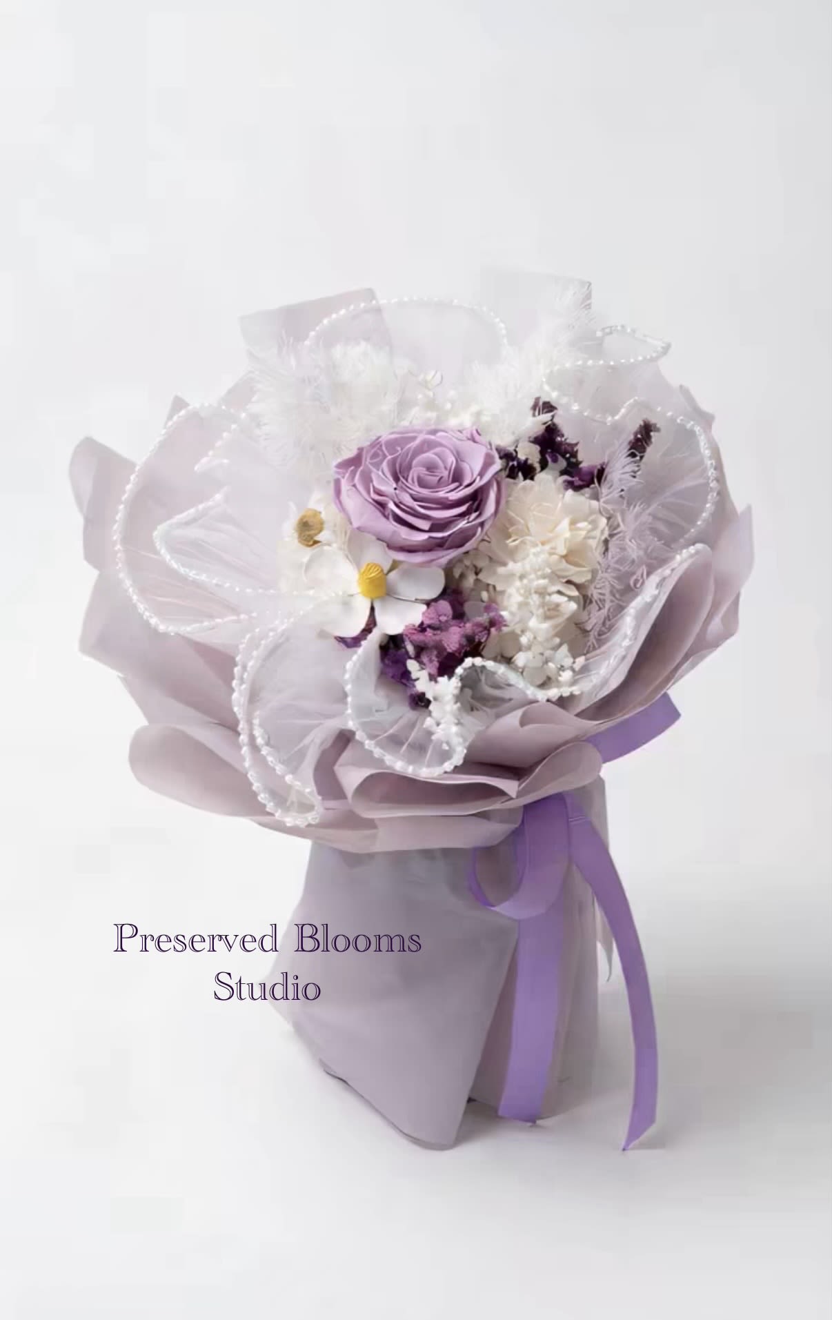 Hand-tied bouquet of lavender and white flowers wrapped in lilac paper with pearl accents and ribbon.
