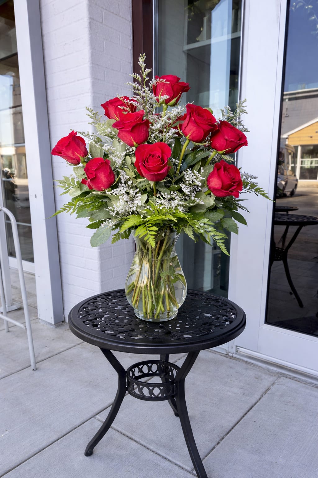 Arrangement of long-stem red roses with white filler flowers in a clear glass vase on a black metal table.