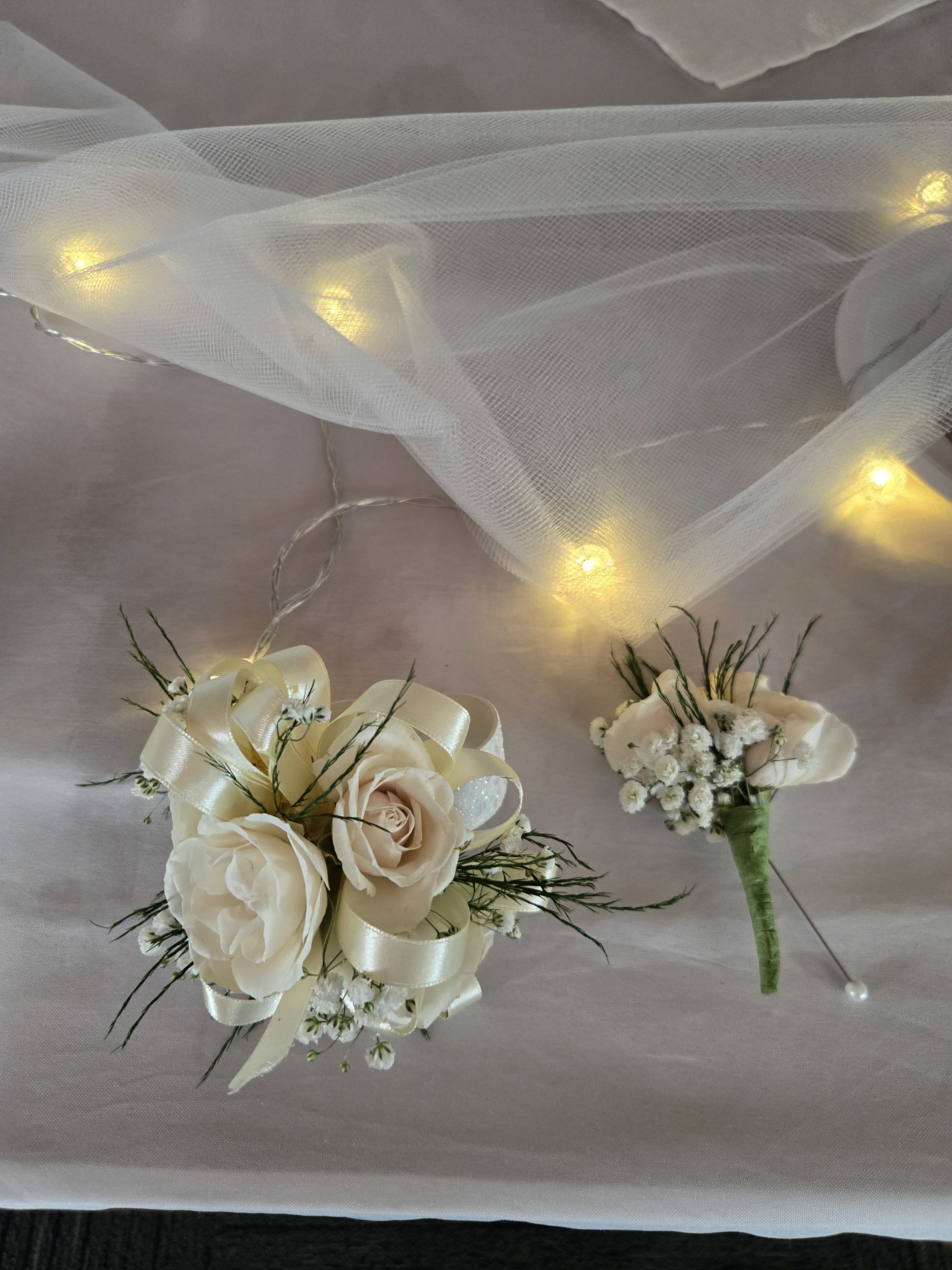 Ivory rose wrist corsage and matching boutonniere with ribbon on a draped cloth with soft lights.