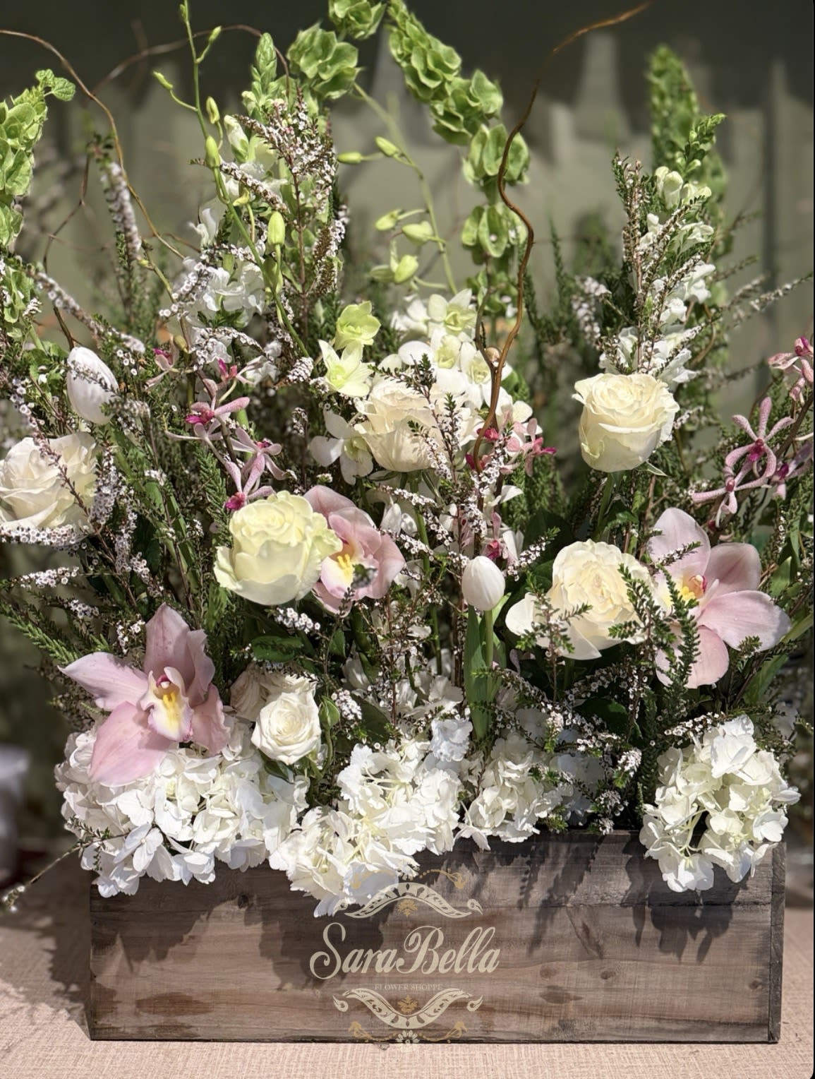 IVORY GARDEN - This is a lush, garden-style floral arrangement with a soft, romantic aesthetic. It’s designed in a rustic wooden boxThe composition is airy and organic rather than tightly structured. You’ve got a mix of: Cream and white roses as the focal flowers Pale pink orchids adding elegance and a delicate pop of color White hydrangeas clustered at the base for fullness Wax flowers and small filler blooms adding texture Greenery and branching stems that create height and movement