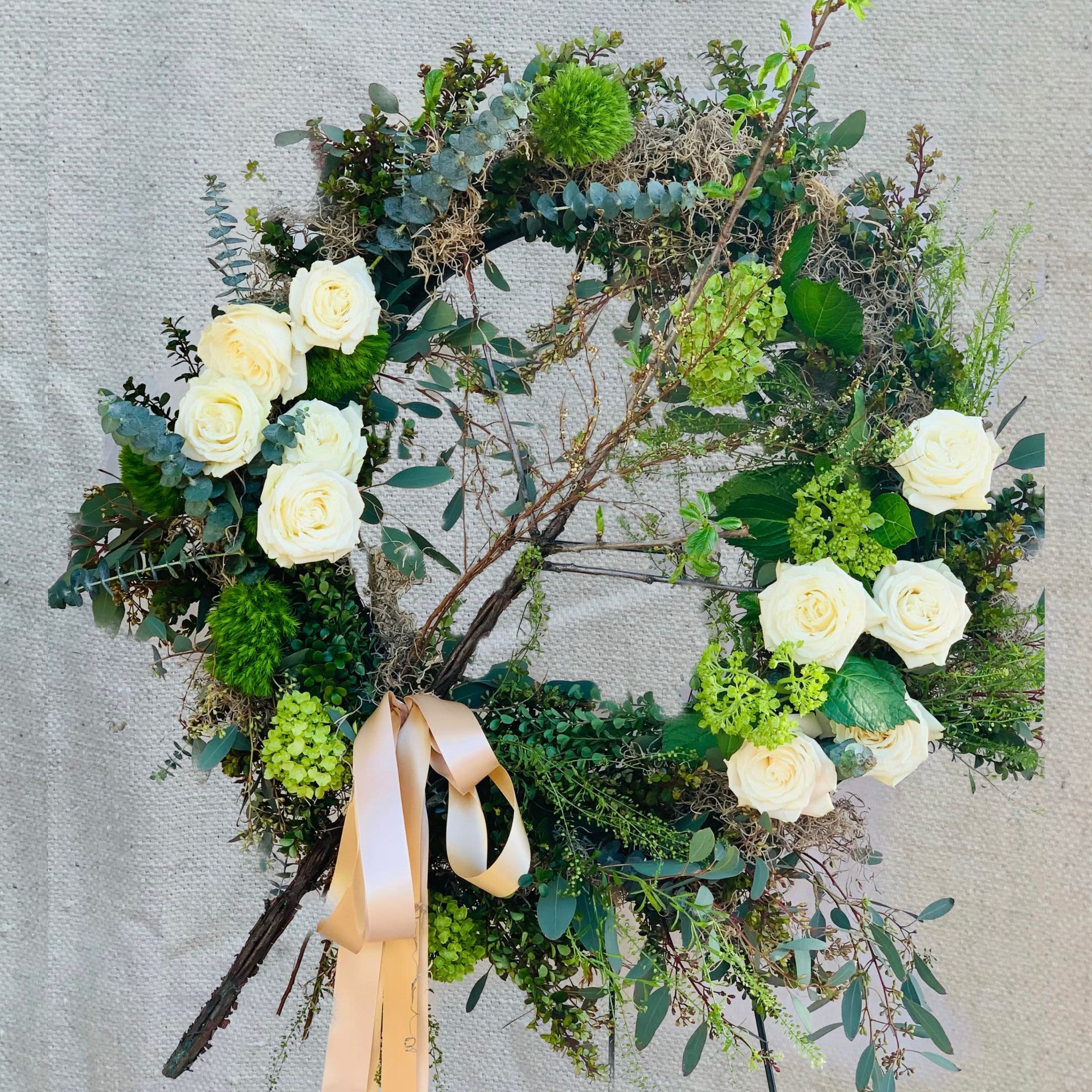 Enlightenment - A naturally elegant sympathy wreath featuring white garden roses and vibrant green blooms set on a rustic grapevine base with lush eucalyptus and textural branches. This organic tribute combines fragrant white garden roses, green chrysanthemums, green hydrangea, dianthus, eucalyptus, and natural branches, creating a peaceful expression of eternal life and natural beauty.  Specific florals can be included or removed and colored themes are available. Each wreath includes a 5' wire easel and is expertly crafted with the freshest blooms and greenery.