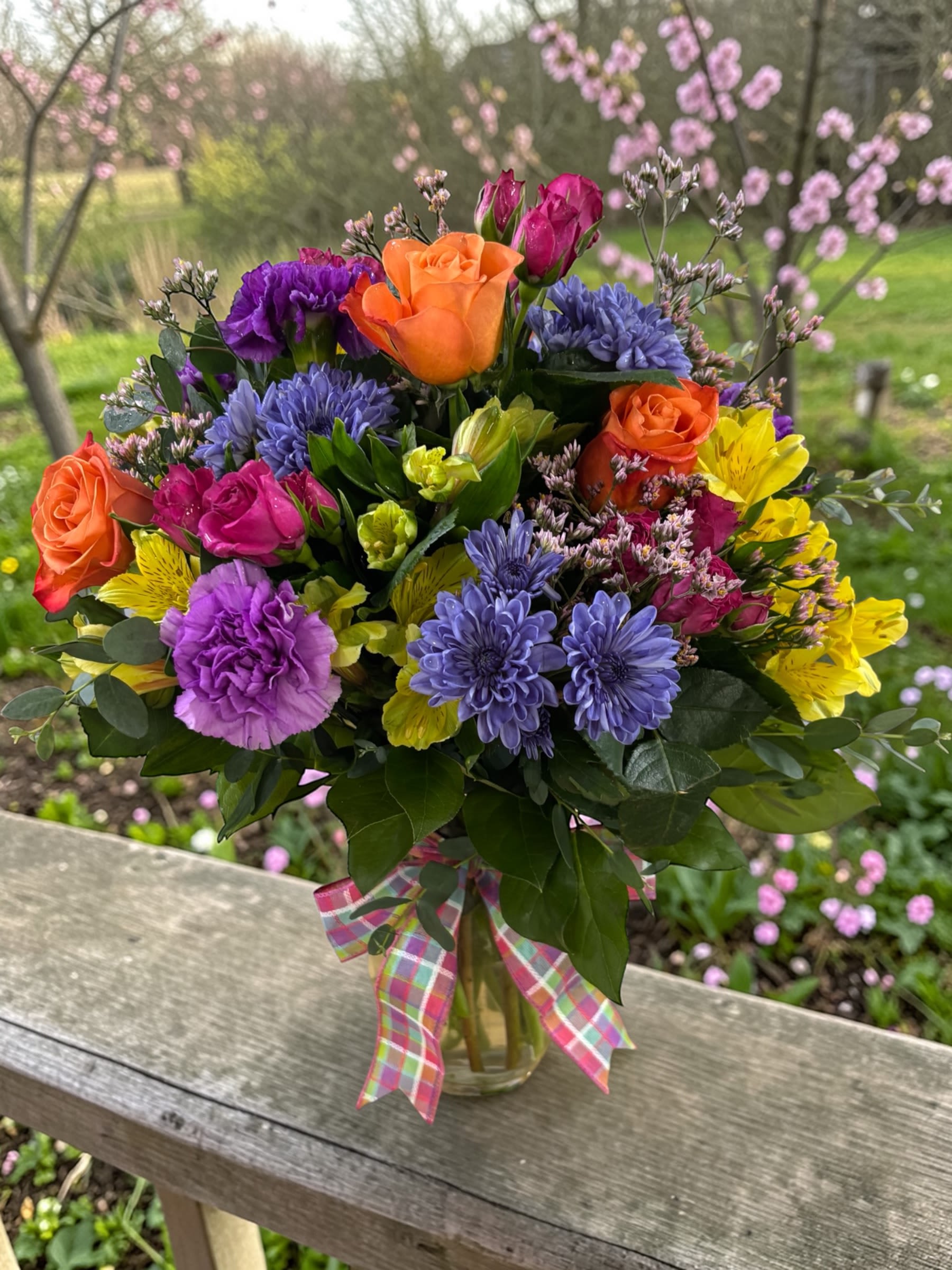 Spring Explosion  - Color lovers, this one’s for you. Bursting with vibrant orange roses, bold fuchsia spray roses, sunny yellow alstroemeria, rich purple carnations, and lavender mums, this arrangement delivers bright, layered color in a compact, cheerful design.  The palette does the work here—energetic, balanced, and full of personality—creating a piece that instantly lifts the space without feeling overdone.  Designed in a smaller size for easy pickup, this version keeps the same bold look and feel while offering a more accessible option.  A perfect choice for birthdays, thank-you gifts, thinking of you, or anyone who could use a little extra color in their day.  Each arrangement is created with the freshest stems available. Varieties may vary slightly based on availability while maintaining the same vibrant color palette.  Available for pickup or local flower delivery through Aspen Tower Florist, serving Greensburg and Shelbyville along with Rushville, Batesville, Westport, Oldenburg, St. Paul, Waldron, Milroy, and Hope.