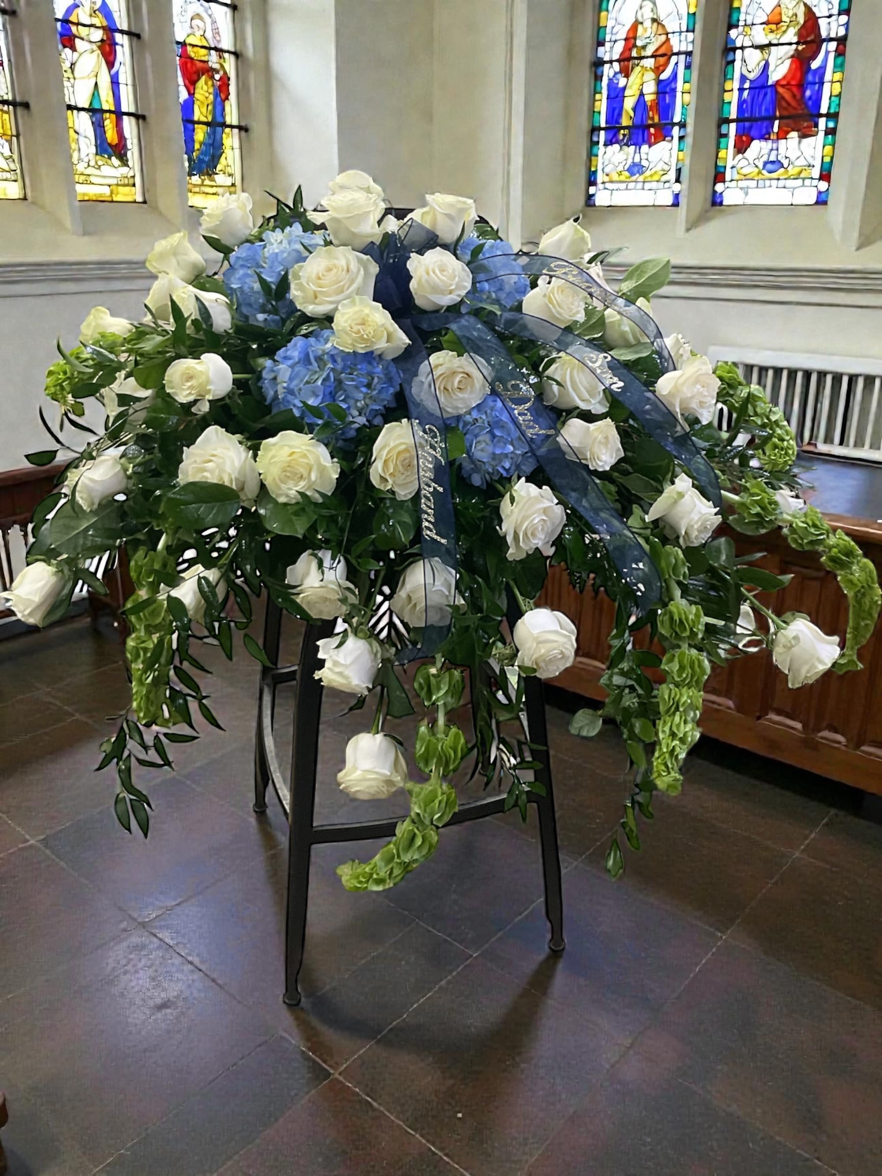 Canvas of Heaven - Soft, serene, and quietly elegant, Canvas of Heaven is a casket spray created to honor a life with grace, faith, and remembrance. Premium white roses form the heart of the design, accented by sky-blue hydrangea and flowing bells of Ireland that cascade naturally across the casket. The result is a peaceful tribute filled with movement, texture, and understated beauty.  This design was originally created for a family served through Todd Funeral Centre &amp; Crematory, 1600 S State Road 3, Rushville, IN 46173. Families may learn more about Todd Funeral Centre &amp; Crematory at www.toddfuneralcentre.com.  Customers can view additional examples of our custom casket sprays in the Casket Spray section of our website, including real photographs and videos of funeral tributes created by Aspen Tower Florist.  A meaningful choice for funeral services, memorial services, and celebrations of life, Canvas of Heaven offers a timeless combination of white roses, blue hydrangea, and lush greenery designed to provide comfort, beauty, and a lasting expression of love.