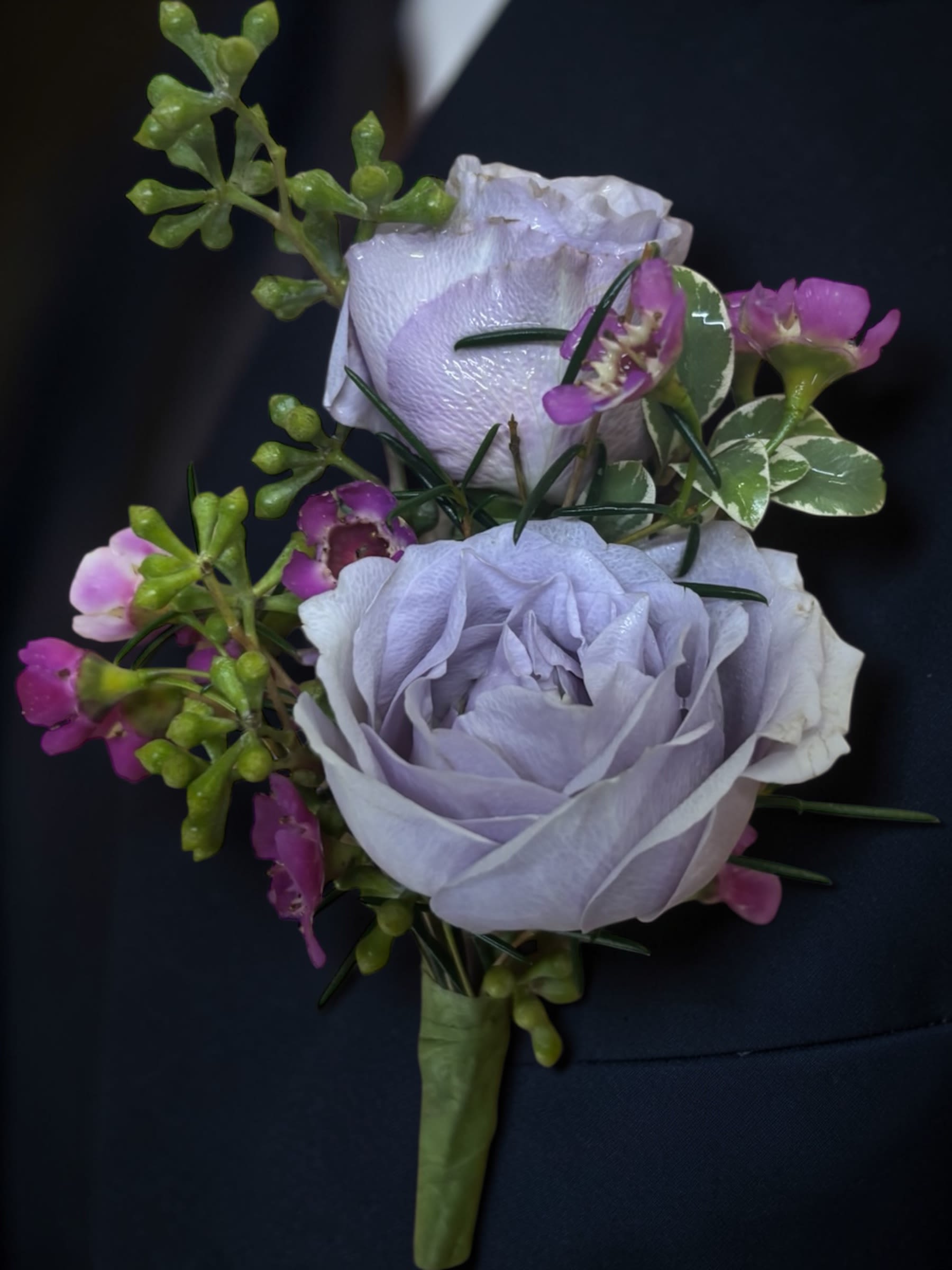 Lavender Skies Wedding Boutonniere  - Lavender Skies Boutonnière features lavender spray roses accented with complementary flowers, variegated greenery, and natural textures for a timeless wedding look filled with romance and elegance. Designed to coordinate beautifully with the Lavender Skies wedding collection, this boutonnière brings together soft lavender tones, fresh greenery, and delicate floral accents in a refined design that complements the bridal bouquet, bridesmaid bouquets, centerpieces, corsages, and ceremony flowers.  The combination of lavender roses and carefully selected accents creates dimension, texture, and visual interest while maintaining a clean, classic appearance. Soft color and natural movement make this boutonnière an elegant choice for grooms, groomsmen, fathers, grandfathers, ushers, officiants, and special family members.  Designed to complement both traditional and garden-inspired wedding styles, this boutonnière provides a polished finishing touch while remaining comfortable and lightweight to wear throughout the day. The soft lavender color palette adds a romantic element without overwhelming formal attire, creating a coordinated look across the entire wedding party.  Flowers and accents may vary based on seasonal availability while maintaining the overall style, color palette, quality, and feel of the design. A beautiful finishing touch designed to bring elegance, romance, and coordinated floral beauty to your wedding day.