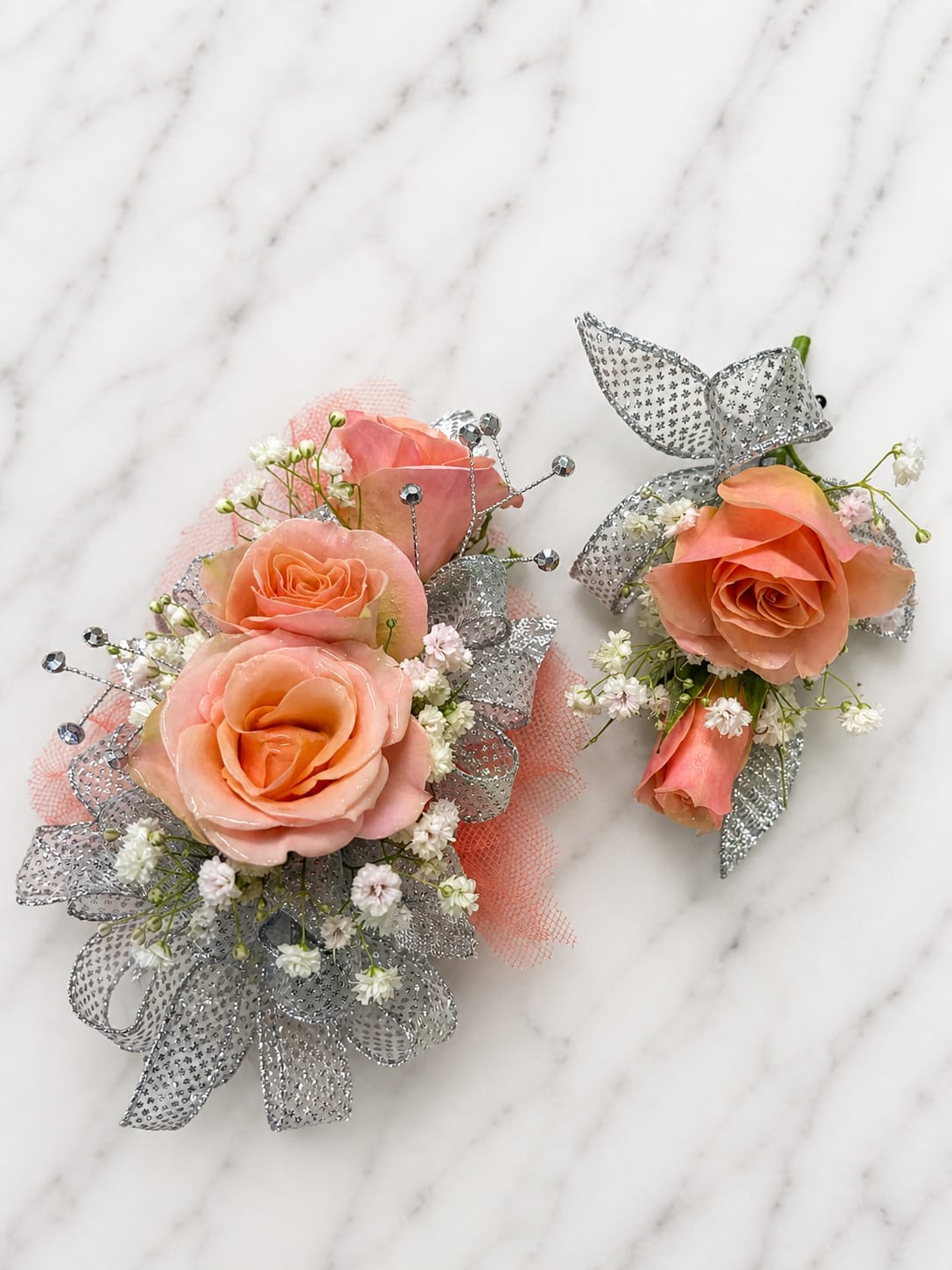 Peach Shimmer Formal Set - MAGNETIC BOUTONNIERE WARNING: CONTAINS STRONG MAGNETS. DO NOT USE IF YOU HAVE A PACEMAKER, DEFIBRILLATOR, INSULIN PUMP, COCHLEAR IMPLANT, NEUROSTIMULATOR, OR OTHER IMPLANTED MEDICAL DEVICE. KEEP AWAY FROM ELECTRONICS, CREDIT CARDS, HOTEL KEY CARDS, AND CHILDREN.  Peach Shimmer  Soft peach roses, sparkling silver ribbon, delicate filler flowers, and shimmering accents come together in Peach Shimmer, an elegant corsage and boutonniere collection designed for prom, homecoming, dances, banquets, weddings, and formal events. The combination of warm peach tones and cool silver accents creates a timeless look that is both graceful and eye-catching.  Standard includes a wrist corsage with 3 spray roses and a matching magnetic boutonniere with 2 roses. Deluxe includes 4 spray roses in the corsage. Premium includes 5 spray roses for additional fullness and flower coverage. All boutonnieres include 2 roses.  Perfect for prom flowers, wrist corsages, magnetic boutonnieres, corsage and boutonniere sets, homecoming flowers, dance flowers, and formal event flowers.  Because each corsage and boutonniere is individually designed, no two sets will be exactly alike. Ribbon, filler flowers, bracelets, decorative accents, and other design elements may vary based on availability. While individual details may differ, the overall color palette, roses and/or flowers, style, and appearance of the Peach Shimmer collection will be maintained.