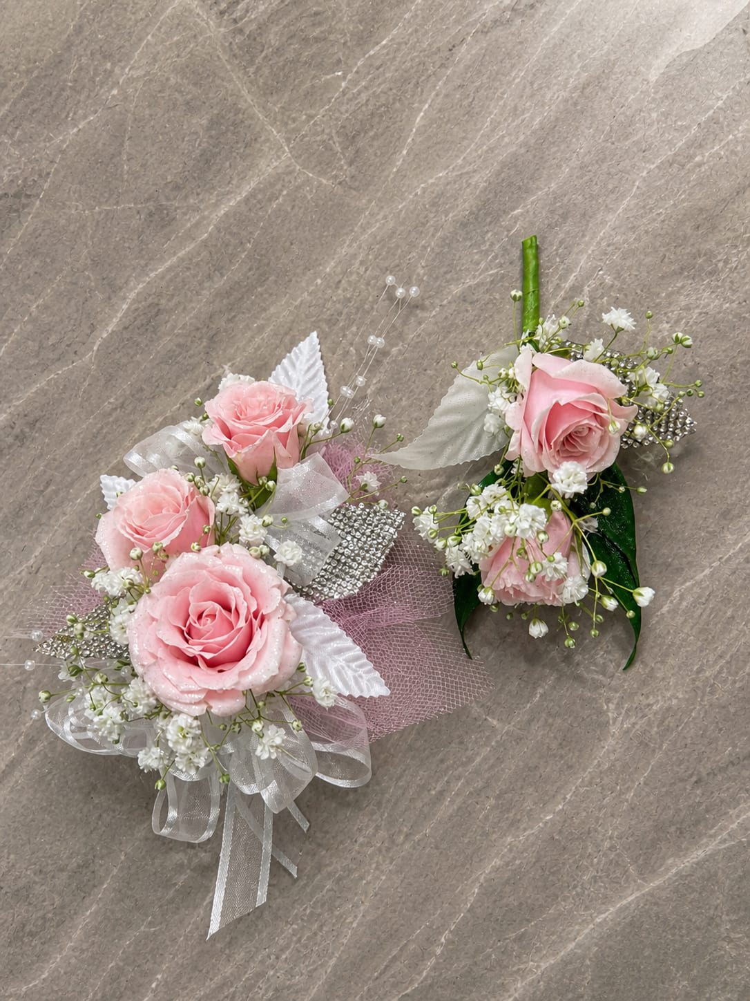 Pink Ice Formal Set - MAGNETIC BOUTONNIERE WARNING: CONTAINS STRONG MAGNETS. DO NOT USE IF YOU HAVE A PACEMAKER, DEFIBRILLATOR, INSULIN PUMP, COCHLEAR IMPLANT, NEUROSTIMULATOR, OR OTHER IMPLANTED MEDICAL DEVICE. KEEP AWAY FROM ELECTRONICS, CREDIT CARDS, HOTEL KEY CARDS, AND CHILDREN.  Pink Ice  Soft pink roses, crisp white accents, sparkling rhinestones, pearl details, and delicate filler flowers come together in Pink Ice, an elegant corsage and boutonniere collection designed for prom, homecoming, dances, banquets, weddings, and formal events. The combination of blush pink, white, silver sparkle, and pearl accents creates a fresh, graceful look with just the right amount of shimmer.  Standard includes a wrist corsage with 3 spray roses and a matching magnetic boutonniere with 2 roses. Deluxe includes 4 spray roses in the corsage. Premium includes 5 spray roses for additional fullness and flower coverage. All boutonnieres include 2 roses.  Perfect for prom flowers, wrist corsages, magnetic boutonnieres, corsage and boutonniere sets, homecoming flowers, dance flowers, and formal event flowers.  Because each corsage and boutonniere is individually designed, no two sets will be exactly alike. Ribbon, filler flowers, bracelets, decorative accents, and other design elements may vary based on availability. While individual details may differ, the overall color palette, roses and/or flowers, style, and appearance of the Pink Ice collection will be maintained.