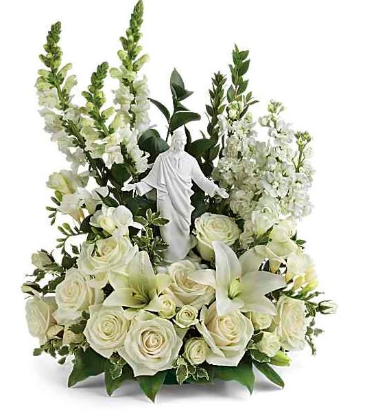 Garden of Serenity Bouquet - White roses and white lilies rise around a small angel figurine in a dense upright arrangement crafted as a standing tribute-perfect for a sympathy arrangement or memorial service. The Garden of Serenity bouquet offers a devotional, comforting presence that helps the person you're thinking of feel honored and remembered during a difficult moment.  Clustered white roses and lilies are offset by tall white spires and dark green foliage to create a formal, full silhouette; layered blooms give the piece texture while the contrasting greenery keeps the palette serene and restful. A delicate angel figurine sits at the center, making this arrangement appropriate for funerals, memorials, or as a sympathy gift to convey peace and consolation.  Designed one-sided for presentation at a service, this bouquet reads as a carefully composed tribute rather than a casual bouquet. If any listed flowers are unavailable, local florists will substitute with the freshest comparable blooms to preserve the overall look and thoughtful intent.