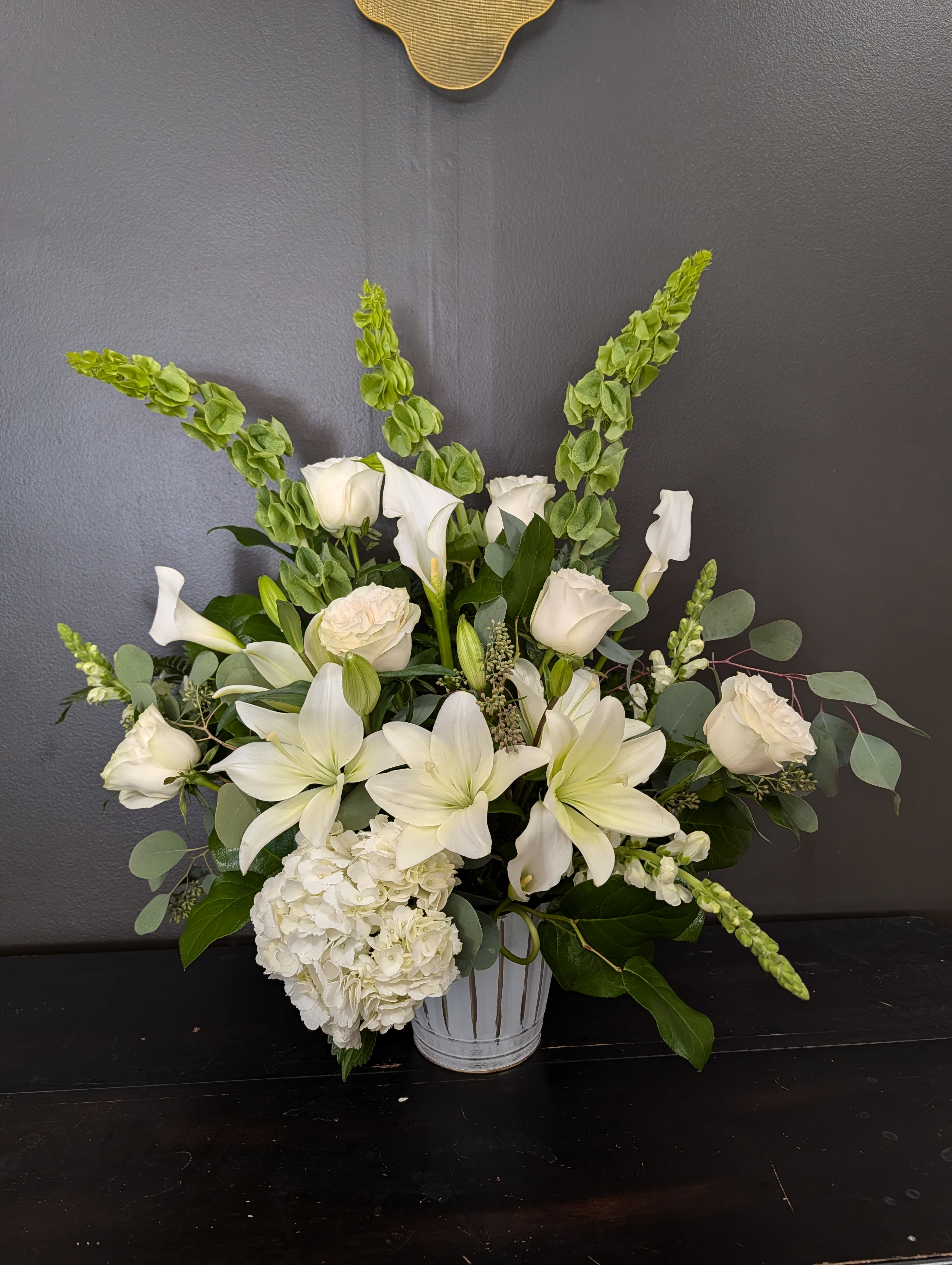 Luck Be A Lady - An all-white funeral basket featuring calla lilies, roses, delphinium orchids, and more framed by a backdrop of large leaves.