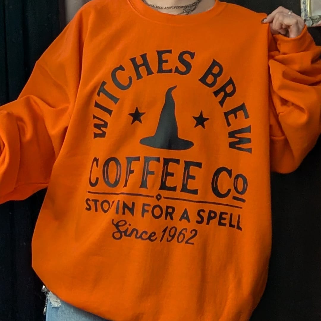 Orange sweatshirt with black witch-themed coffee shop text and hat graphic