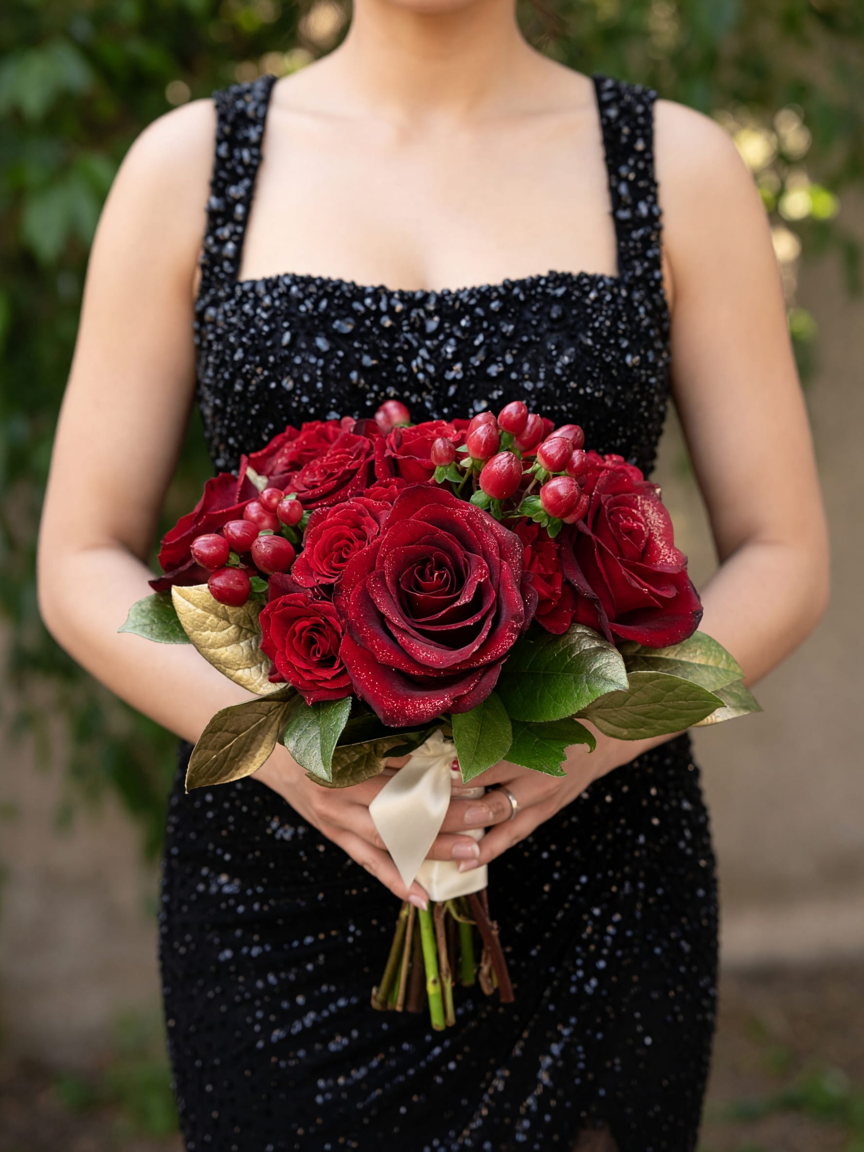 Red and Gold Nosegay  - Tightly clustered deep red roses form the heart of this romantic flower arrangement, a red and gold nosegay designed for anniversaries, Valentine's Day, or any moment you want someone to feel truly cherished. The compact, rounded shape reads formal and intentional-she'll feel noticed and celebrated the moment she sees the rich crimson blooms against metallic green-gold foliage.  Small red berry-like accents add texture while broad, green-gold leaves give the bouquet a luminous, festive edge. Stems are bound with a pale satin ribbon for a polished finish; the arrangement reads medium-small and is meant to be held or presented as a classic hand-tied bouquet. Ideal for gifting to a partner or close loved one, it delivers a confident, romantic message with minimal fuss-simple care is to keep the stems in fresh water and trim them slightly when you rearrange the display.