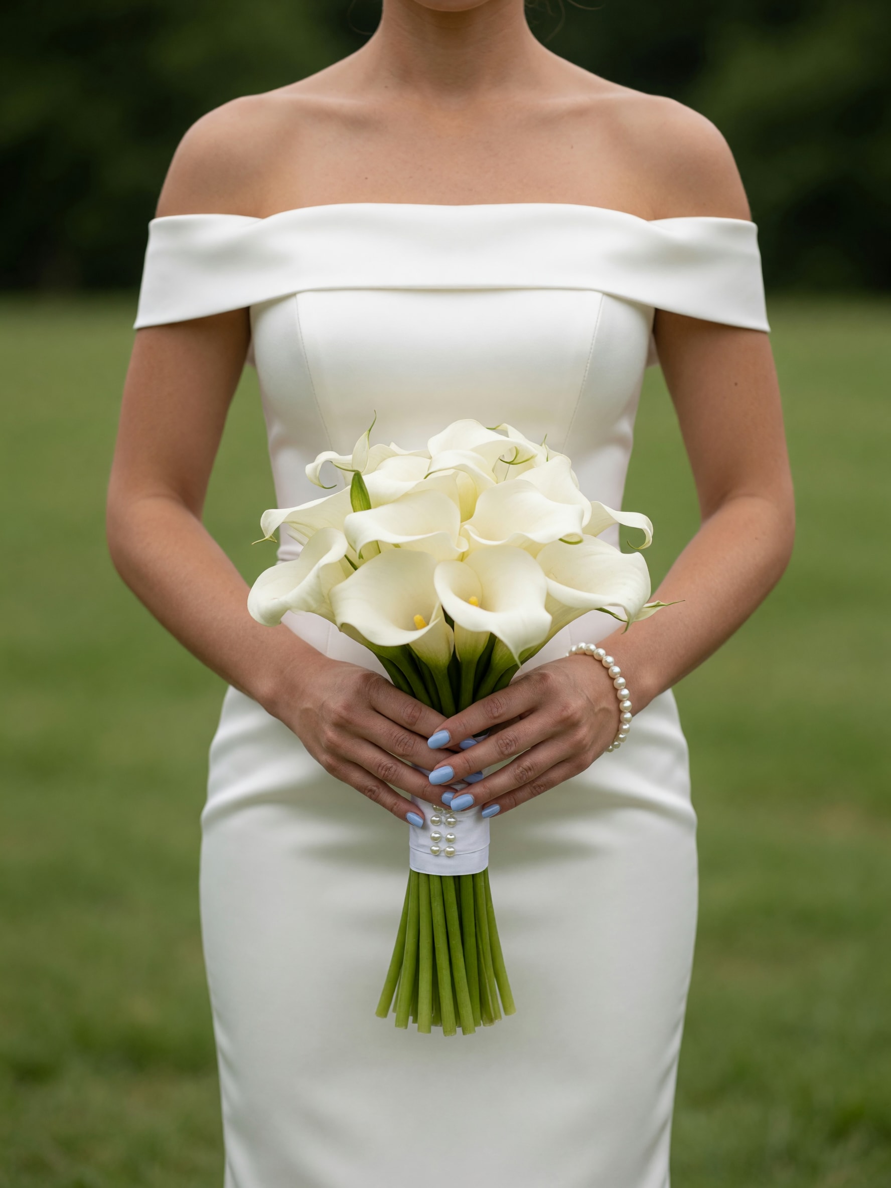 Calla Lily Bouquet - Pure white calla lilies arranged in a tight, sculptural cluster-this wedding bouquet feels both modern and timeless. As a bridal bouquet or an elegant choice for an anniversary or formal event, the clean white and green palette reads classic; the smooth, rolled petals create a refined silhouette that makes the person you're gifting feel composed and celebrated.  The arrangement is compact with a rounded top of long-stemmed white calla lilies bound into a narrow handle. Finished with white satin ribbon and delicate pearl embellishments, the simple wrapping keeps the focus on the lilies' sleek form and smooth texture. The stems extend straight downward for a streamlined, architectural look ideal for a ceremony or portrait photos.  Perfect for a bride seeking minimalist sophistication or anyone marking a significant milestone, this bouquet sends a message of purity and graceful confidence. Hand-delivered ready to carry; soak the stems briefly in water before arranging in a vase to extend freshness if kept after the event.