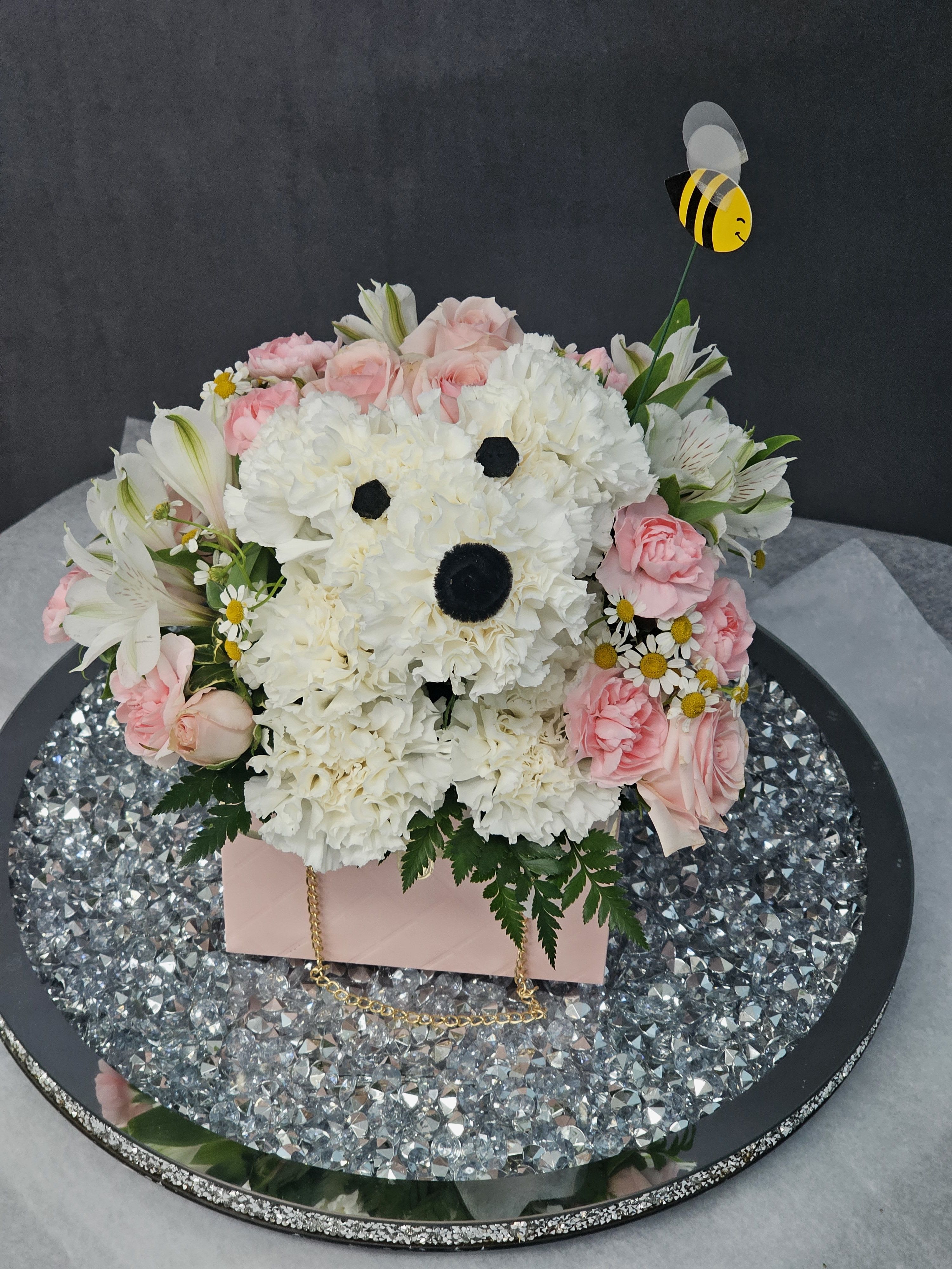 Petal Pup - Dense pastel arrangement of white carnations, pink spray roses, white alstroemeria, and pink mini carnations in a pink gift box with bee topper .