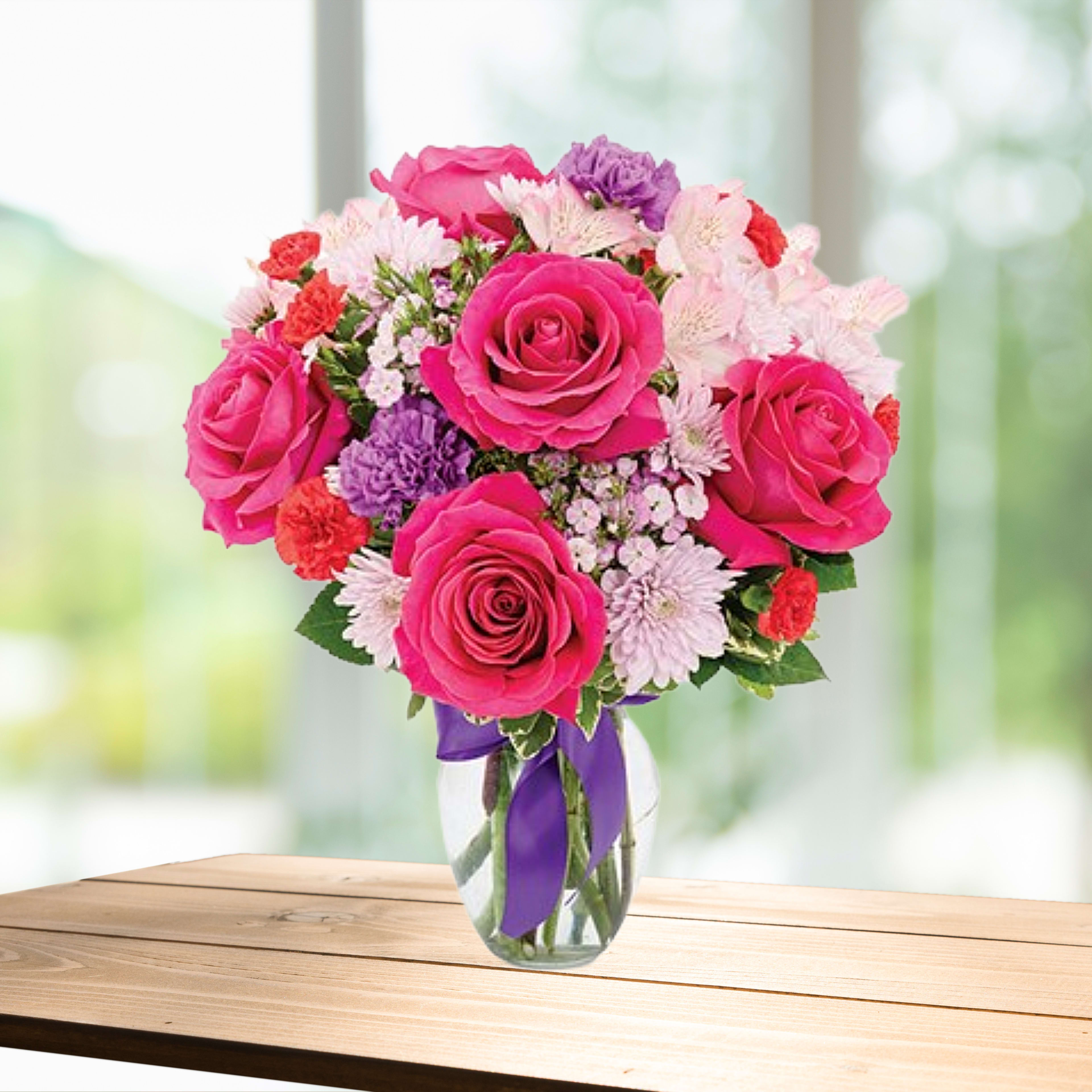 Pink roses and mixed flowers in a clear glass vase with a purple ribbon