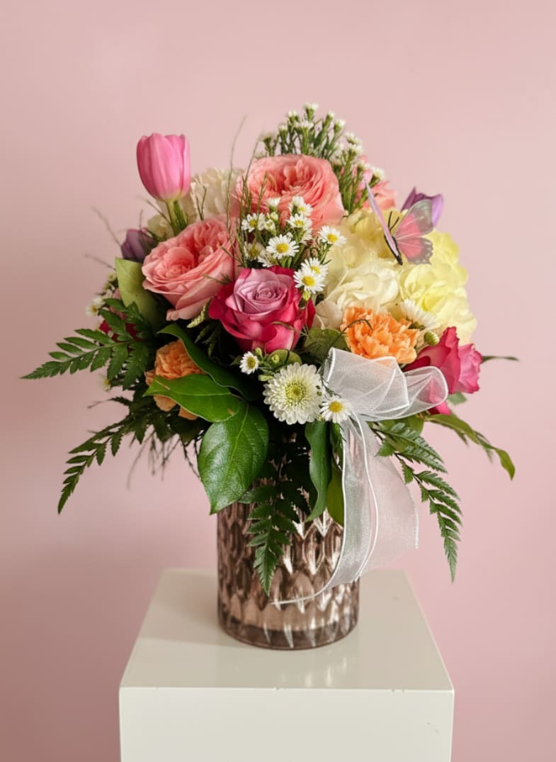 Queen Bee - Pink roses and soft pastels take center stage in this rounded, full arrangement crafted to make someone feel truly celebrated. Queen Bee is a flower arrangement for anniversaries, birthdays, or romantic moments-its mix of pink roses, white hydrangeas, purple tulips, orange and peach carnations, pale yellow blooms, and petite white daisy-like filler flowers reads like spring in a vase while remaining appropriate year-round. A sheer white ribbon and a delicate butterfly decoration give it an unmistakably gift-worthy finish that will brighten the recipient's day.  Arranged in a textured glass vase that adds a reflective, polished pedestal presentation, the bouquet balances soft feminine tones with a few brighter accents for cheerful contrast. The composition highlights layered textures-velvety roses, crisp hydrangea clusters, smooth tulip petals, and ruffled carnations-so it looks full from every angle and displays beautifully on a table or mantle.  Ideal for birthdays, anniversaries, or love-and-romance moments, Queen Bee is the kind of bouquet that makes the person you're thinking of feel seen and cherished. For best enjoyment, keep the blooms in fresh water, out of direct sun, and trim stems every few days; the arrangement arrives ready to display and is available for same-week delivery.