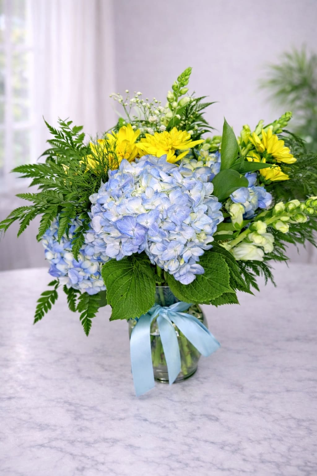 Blue hydrangeas and yellow daisies in a glass vase with a blue ribbon.