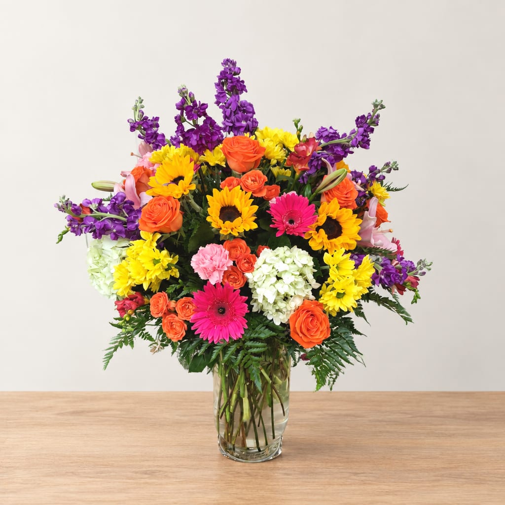 Colorful mixed bouquet in a clear glass vase with sunflowers, roses, and gerbera daisies