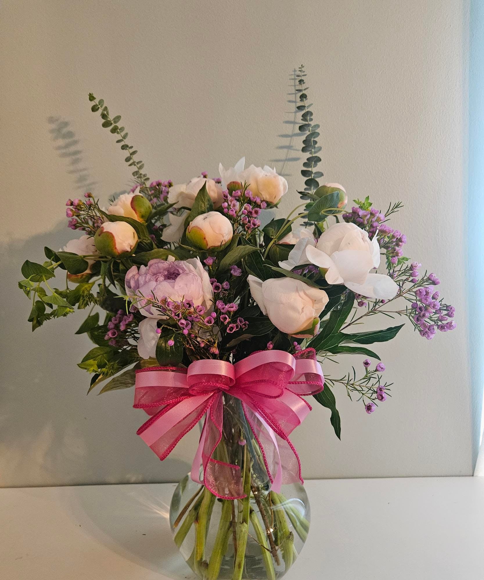 "Great Love Forever" - Soft pink and white peonies take center stage in this romantic flower arrangement, making it an ideal anniversary bouquet or Mother's Day gift that reads as tender and timeless. The rounded cluster is accented with airy purple filler and tall eucalyptus stems that add graceful movement-perfect for saying "I'm thinking of you" on birthdays, get-well days, or simply just because.  Set in a clear glass vase wrapped with layered pink ribbon, the presentation is polished and gift-ready for weddings, housewarmings, or sympathy and funeral tributes where a gentle, comforting touch is needed. The palette of pink, white, green and hints of purple creates a soft, seasonal look that works year-round-from spring and summer celebrations to autumn and winter holidays-while the mix of full peony blooms and delicate sprigs gives appealing texture and scent. Care: keep vase water fresh and trim stems every few days to extend bloom life.