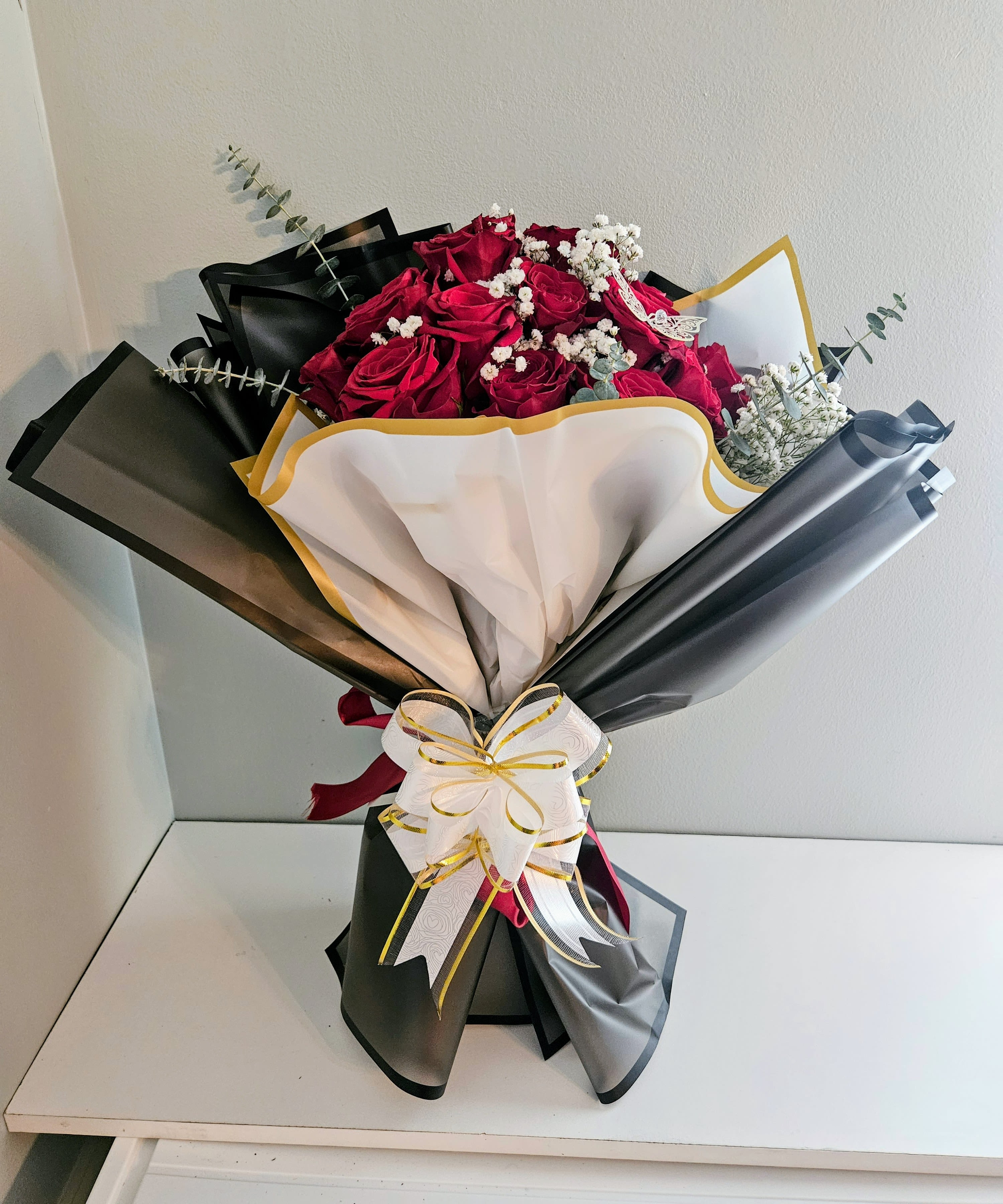 "Red Royal Blossom" - Deep, velvet-red roses gathered tight in a polished gift bouquet-this flower arrangement is a romantic statement perfect for an anniversary or Valentine's Day surprise. The deep red roses create an immediate sense of passion while the small white filler blooms and sprigs of eucalyptus add gentle contrast and fresh texture, so the person you're celebrating will feel truly cherished the moment they see it.  Wrapped in layered black, white, and gold-edged paper with a white ribbon trimmed in gold, the Red Royal Blossom reads as dramatic and elegant; a butterfly pick tucked into the stems gives a delicate, personal touch. The arrangement combines classic roses with subtle greenery and refined hard goods for a high-contrast presentation that photographs beautifully and travels well.  Choose this bouquet when you want to mark a romantic milestone or make someone feel adored and seen. Delivered ready to display, it arrives wrapped and tied-no assembly required-so the recipient can enjoy the visual impact right away; keep stems in water and refresh as needed to extend the life of the roses.