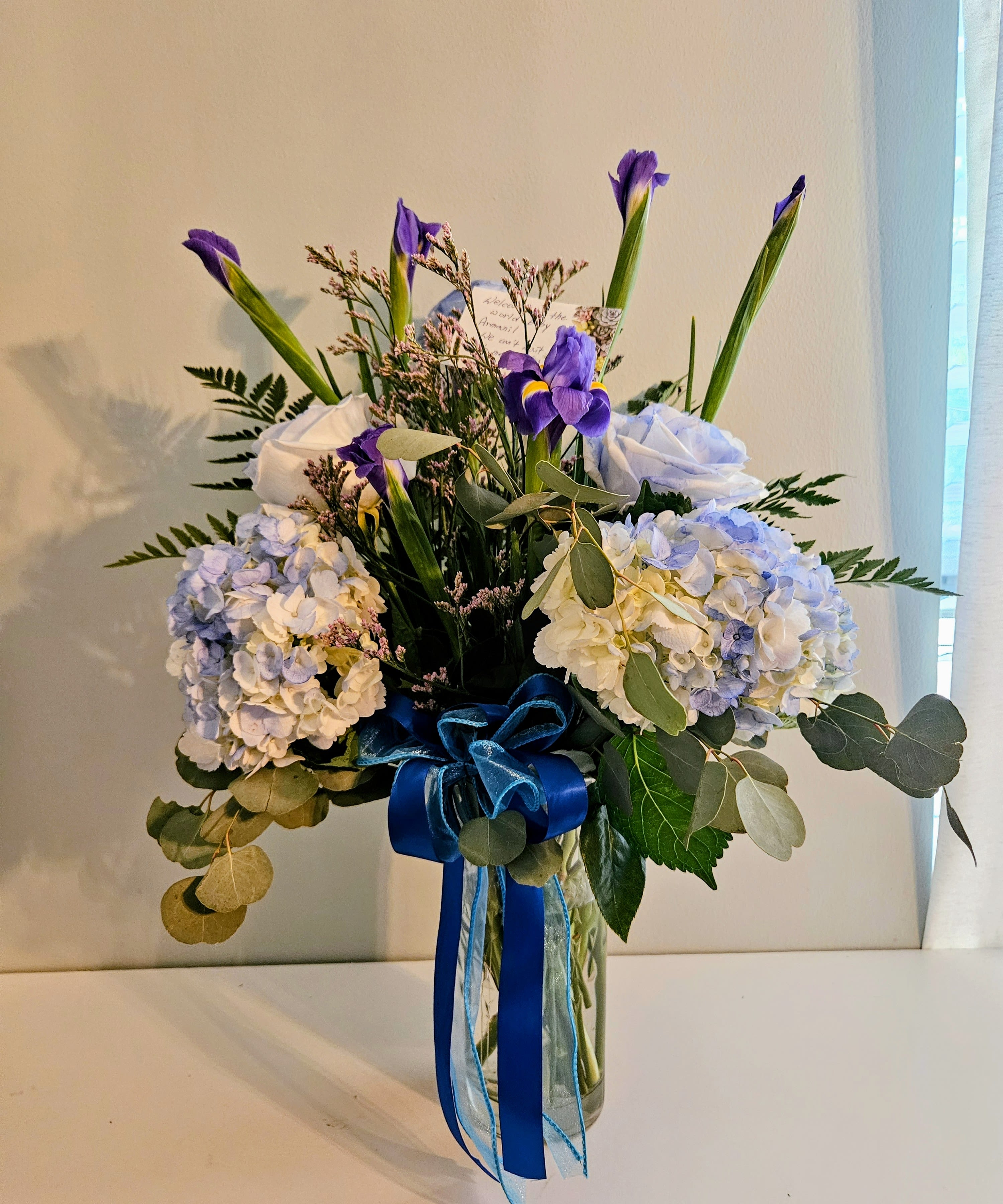 "Blue Light Misty" - Tall purple irises mingle with rounded clusters of pale blue and white hydrangeas in this airy flower arrangement, creating a calm, elegant bouquet perfect as a birthday bouquet, sympathy arrangement, or spring gift. The asymmetrical design lets the irises reach upward while soft hydrangea blooms form a cloud of cool blues and whites, so the person you're thinking of will feel noticed and comforted by its quiet sophistication.  Made with hydrangeas, irises, and a few white roses for contrast, the piece is arranged in a clear glass vase tied with layered blue ribbon for a refined finishing touch. Green foliage and extended stems give the composition an open, natural silhouette; the cool purple, blue, white, and green palette reads formal but approachable, ideal for expressing thanks, offering condolences, or celebrating a seasonal moment.  This is the kind of everyday floral gift that suits many occasions-birthdays, thank-you gestures, sympathy, or a fresh spring surprise-so it's easy to choose when you want to make someone feel truly seen. Care: keep the vase filled with fresh water and trim stems every few days to prolong bloom life.