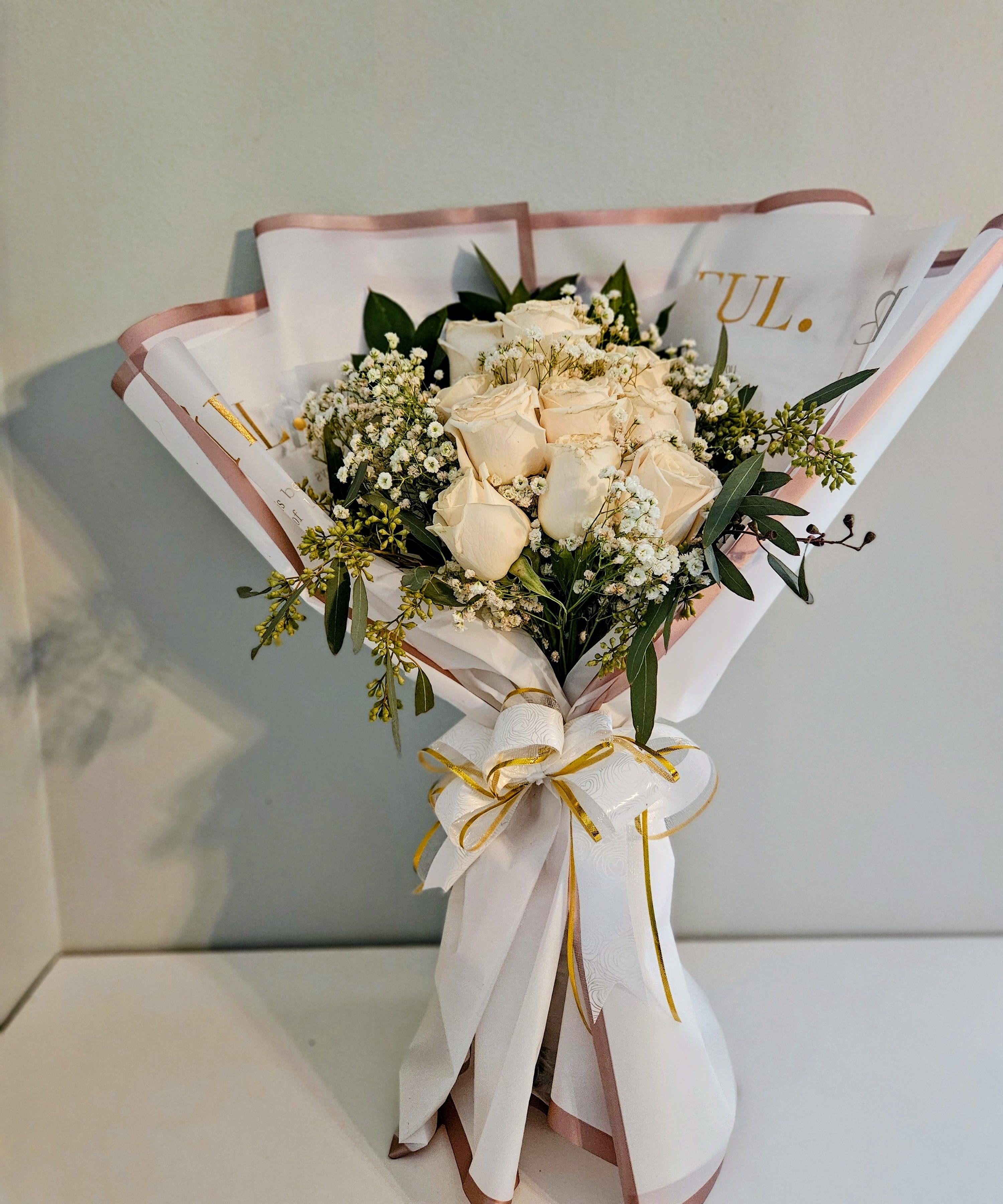 "Ivory Misty" - Creamy roses at the heart of this flower arrangement create a refined, romantic statement-perfect as an anniversary bouquet, birthday flowers, or a thoughtful thank-you gift. The Ivory Misty arrangement pairs those soft cream roses with delicate white filler blooms and fresh green sprigs to give the person you're thinking of a calm, elegant impression that feels both special and quietly sophisticated.  Wrapped in white decorative paper with a blush-pink edge and finished with a layered satin ribbon bow in white and gold, this bouquet reads as polished and gift-ready. Subtle touches of pink and a hint of yellow in the overall palette lift the neutral tones, making it versatile for many recipients; the arrangement arrives ready to display, and its classic colors suit bedside tables, office desks, or a mantel-an easy way to make someone feel genuinely seen.