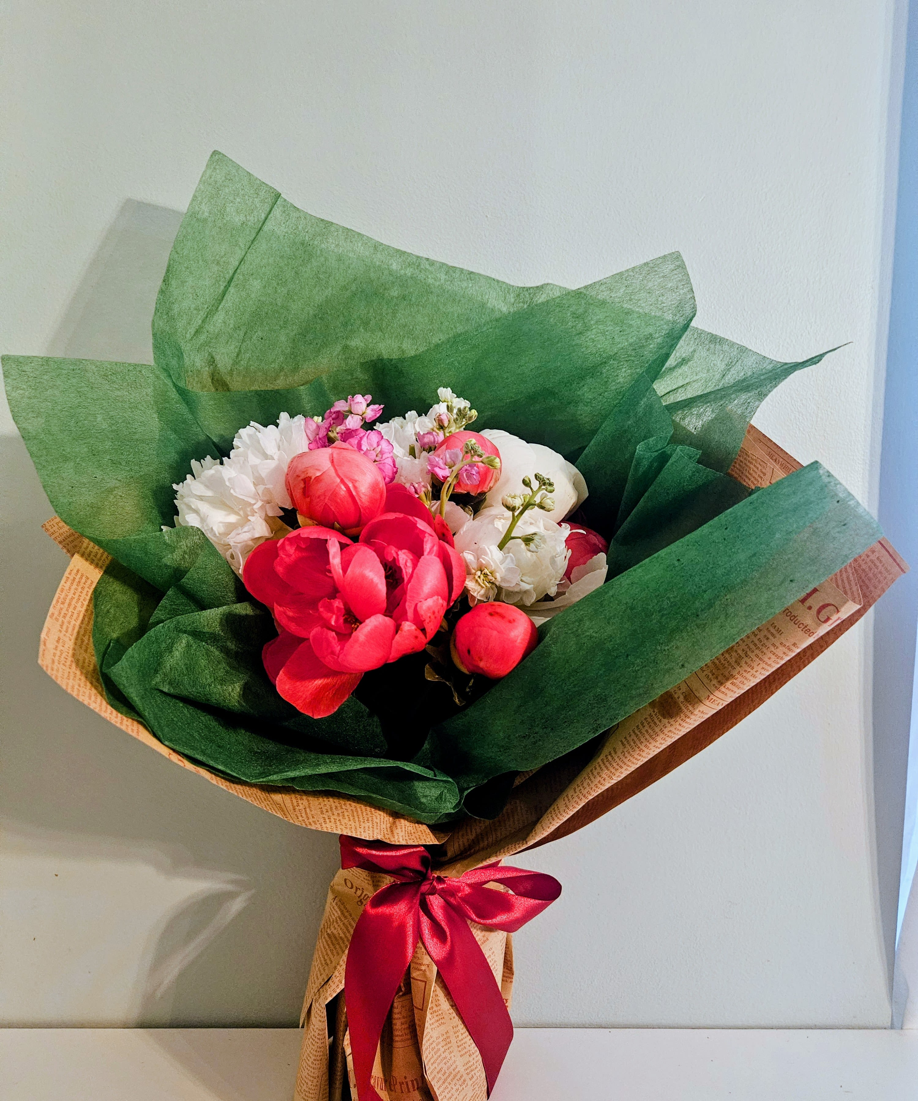 "Peony Party" - A cascade of lush pink peony-like blooms leads this cheerful, romantic flower arrangement-perfect as a birthday bouquet, anniversary bouquet, or a thoughtful thank-you gift. The Peony Party arrangement mixes soft pinks and crisp whites with green accents and a pop of red, so the person you're gifting will feel celebrated the moment they see it.  This bouquet pairs large pink peonies with clusters of white chrysanthemums and upright stems of stock for height and gentle fragrance. Smaller pink accents add texture and depth, all wrapped in deep green floral paper for striking contrast and tied with a red satin ribbon for a polished, gift-ready finish. The layered shapes and varied textures create a full, abundant look that reads as both intimate and joyful.  Send this to someone who deserves to feel truly seen-whether marking a milestone or brightening an ordinary day. Hand-tied and ready to present, the arrangement arrives wrapped and ribboned; refresh stems with water after unwrapping to help the flowers last longer.