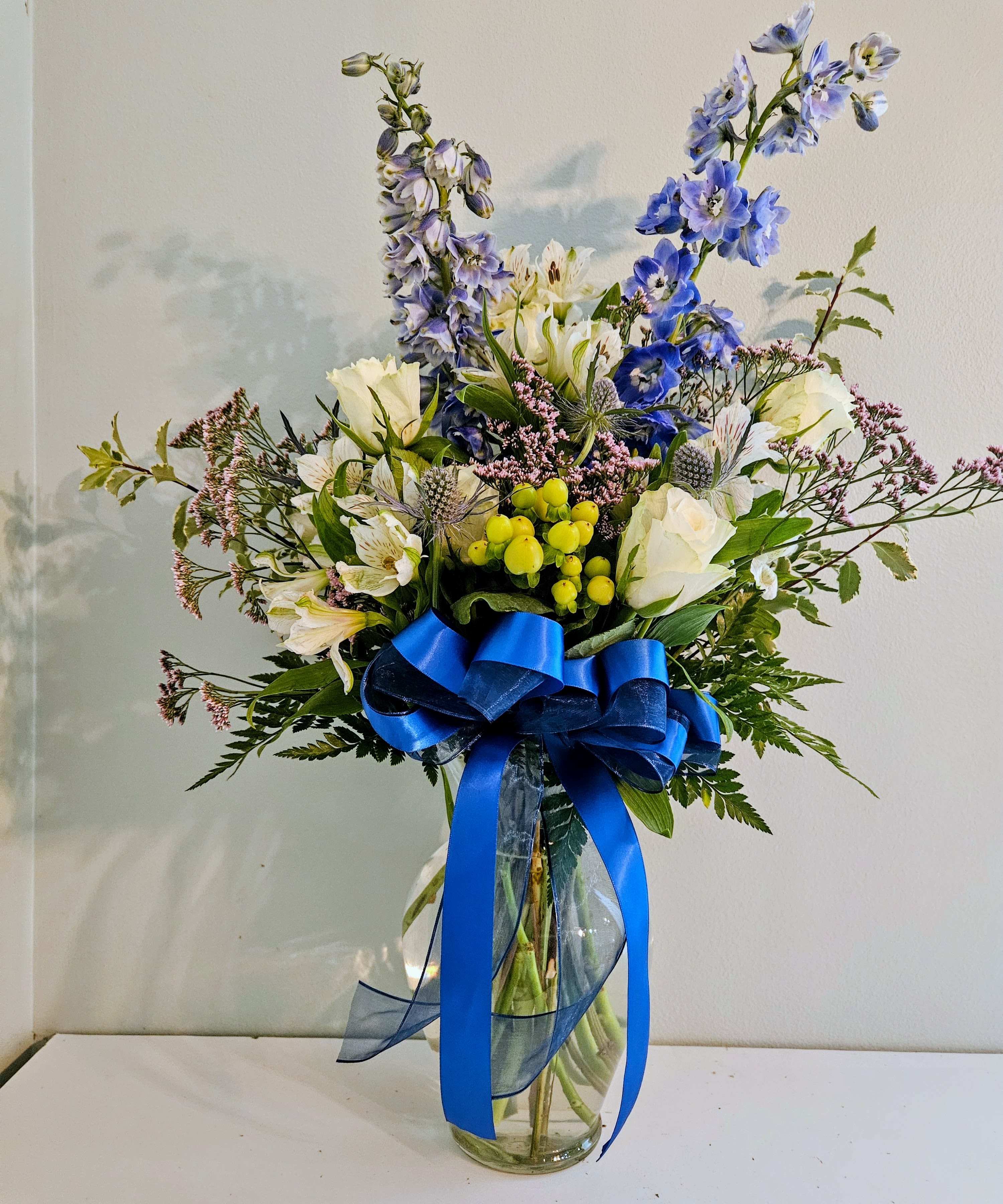 Delight Of Blue - Tall blue delphinium spikes give this flower arrangement a striking vertical presence-the perfect birthday bouquet or thoughtful thank-you gift for someone who loves cool, garden-inspired color. The airy composition reads fresh and elegant, making it equally suited for celebrations, a congratulatory gesture, or to simply let someone know you're thinking of them.  This arrangement pairs upright delphinium with white blooms, soft lavender accents, and roses, accented by small yellow-green berry clusters and delicate filler and fern-like foliage for a loose, garden-style silhouette. It arrives in a clear glass vase tied with a wide royal-blue satin ribbon and a sheer navy wrap, bringing a polished contrast to the natural textures.  Choose this for the person who appreciates refined, cool-toned florals-she'll feel genuinely seen by the arrangement's graceful height and calming palette. The tall vase supports the upright spikes and makes for an impressive display on a console or dining table; standard vase-care-fresh water and a clean vase-will keep blooms looking their best.