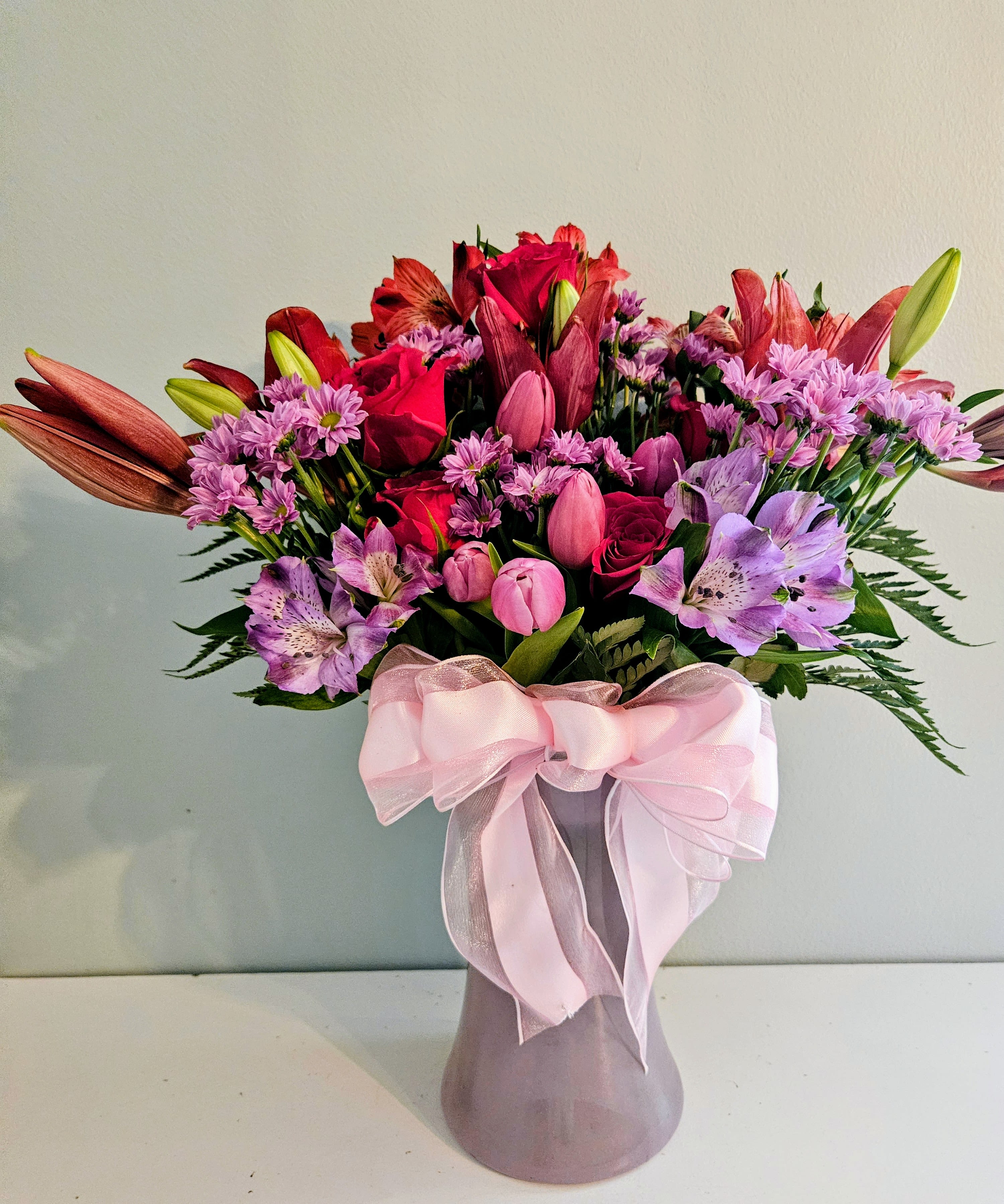 Collaboration With Bright - Hot pink roses and pink tulips take center stage in this vibrant flower arrangement, a birthday and anniversary bouquet that reads as both romantic and joyfully celebratory. Purple alstroemeria and small lavender daisy-like blooms weave through the composition for texture and movement, while dramatic lily blooms and buds lift the silhouette for an elegant, eye-catching profile your recipient will notice from across the room.  Arranged in a frosted glass vase and tied with a wide pink ribbon, the mix of roses, tulips, lilies, alstroemeria, and daisies creates a full, rounded design with strong pink and purple tones softened by creamy whites and grounded by darker greenery at the base. The lilies add height and contrast to the compact clusters of roses and tulips; overall dimensions sit comfortably as a medium-to-large tabletop centerpiece that fills a dining table or entry console.  Choose this bouquet when you want the person you're thinking of to feel special, seen, and celebrated-perfect for birthdays, anniversaries, or simply to brighten someone's day. Vase-arranged and gift-ready, it arrives ready to display; keep stems fresh by changing the water every few days and trimming stems at an angle for longer-lasting blooms.