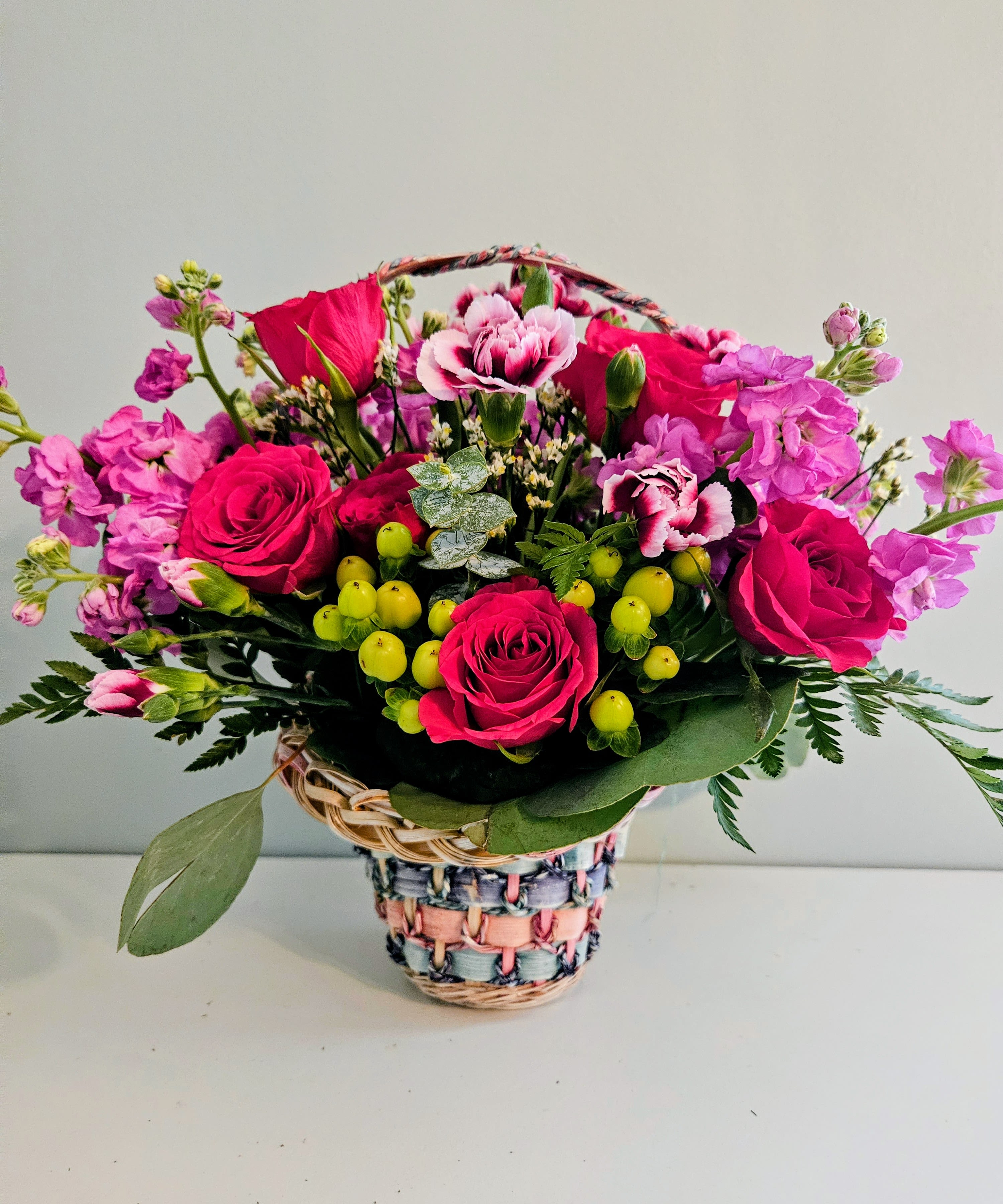 Cute With Pink - Bright magenta and pink roses mingle with pink-and-white carnations in a cheerful rounded basket arrangement - a perfect birthday bouquet or "just because" gift that reads as both playful and heartfelt. The woven basket with a braided handle and multicolored wrap gives the arrangement a handcrafted, sunny look that makes the recipient feel noticed and celebrated, whether you're saying thank you, sending love and romance, or surprising Mom on Mother's Day.  Clusters of small yellow berry-like accents and touches of green foliage add pop and texture against the mixed-color palette of pinks, reds, purples, and yellow highlights. The composition is full and outward-facing, designed to look abundant from every angle and brighten a tabletop or entryway.  This arrangement is sized to be an immediate centerpiece straight from the basket - no vase needed - and arrives ready to enjoy. For best longevity, refresh the floral foam with water every couple of days and keep away from direct heat or drafts so the flowers stay vibrant through the celebration.