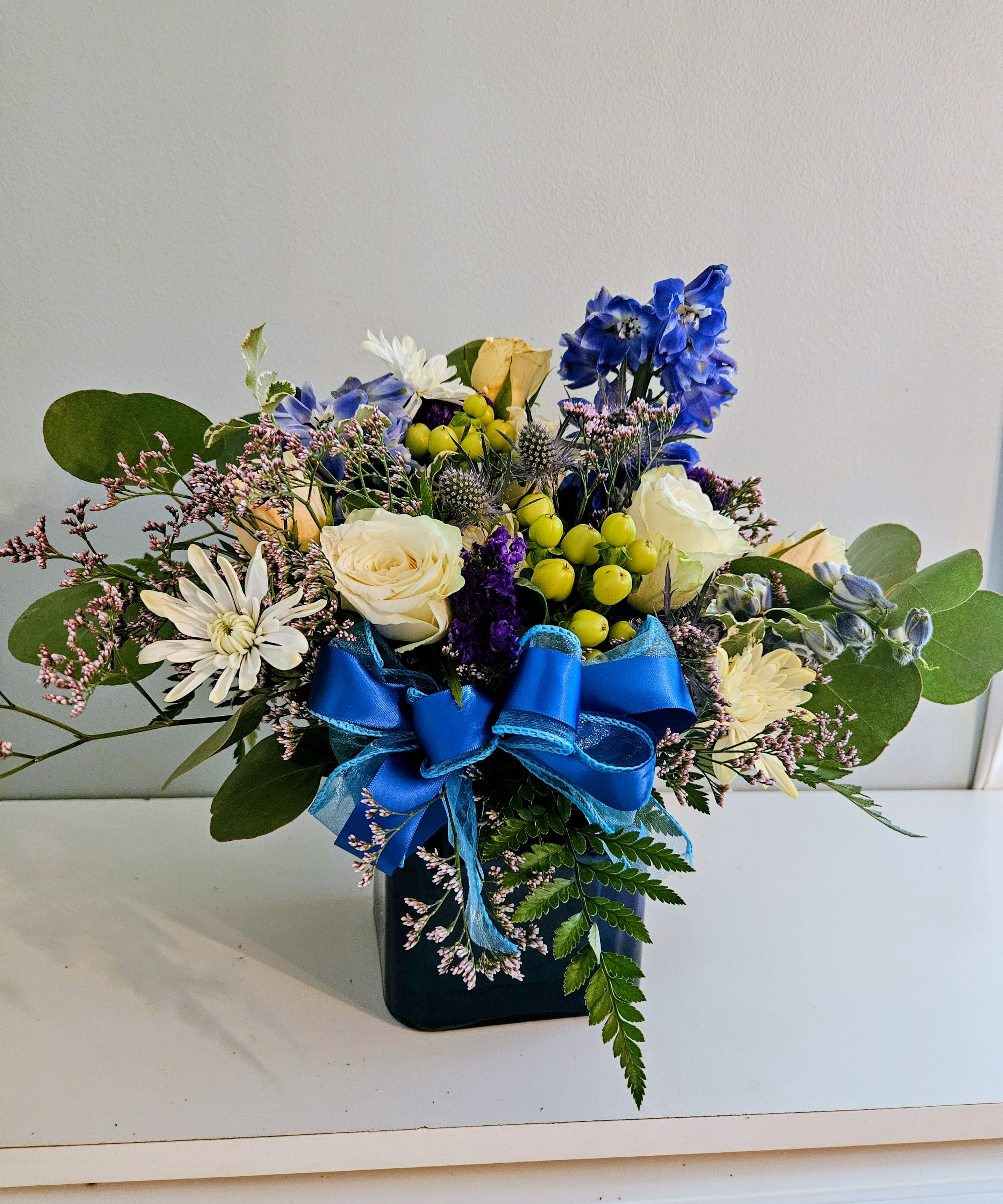 Blue Light - Vivid blue delphinium and cream roses create a fresh, eye-catching centerpiece in this Blue Light flower arrangement - perfect as a birthday bouquet, a cheerful thank-you, or an everyday gift to brighten someone's day. The composition balances bold color with polished presentation so the person you're thinking of will feel truly celebrated the moment it arrives.  Compact and textured, the arrangement pairs cream roses and white daisy-like blooms with small purple filler flowers and bright yellow berry accents for lively contrast. It's arranged in a low black vase and finished with a large blue satin ribbon for a gift-ready look that feels slightly formal yet joyful. The size and close-knit design make it ideal for a table, desk, or bedside display; water the stems and keep in direct sunlight to a minimum for best longevity.