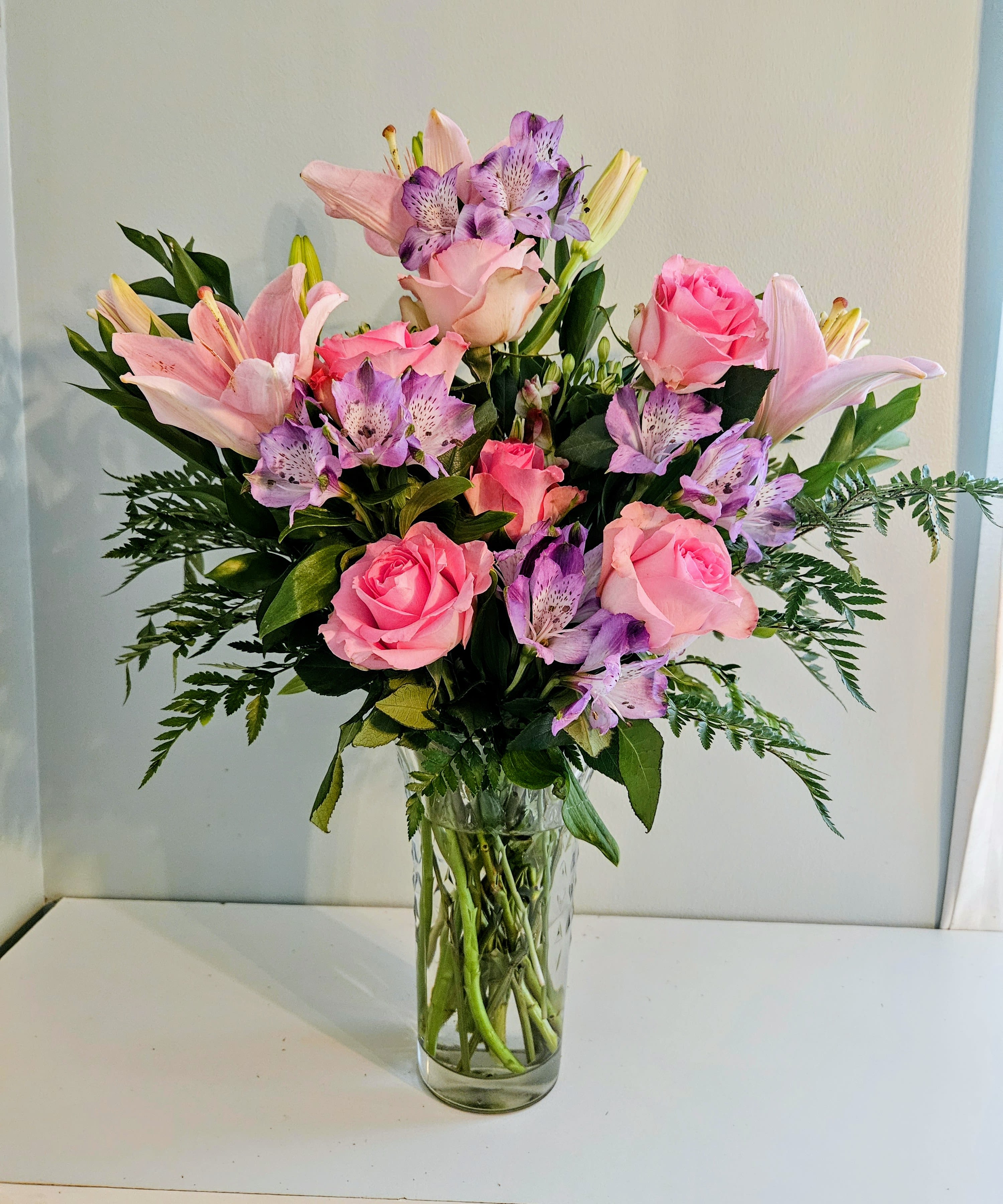 Pink And Purple Swing - Blush roses and pale pink lilies mingle with purple alstroemeria in a full, rounded vase arrangement-perfect as a birthday bouquet, anniversary flowers, or a "thinking of you" gift that feels both romantic and fresh. The soft blend of pink, lavender, white, and green reads like a garden-inspired embrace, so the person you're sending this to will feel noticed and comforted the moment the vase arrives.  Arranged in a clear glass vase, the Pink And Purple Swing features layered blooms with airy, fern-like foliage that extends outward for a balanced, elegant silhouette. Colors include blush and pale pink roses and lilies, bright purple alstroemeria, and touches of white and green for contrast. The design is intentionally full and rounded to give lasting visual impact on a table, desk, or bedside.  This everyday vase arrangement is sized to be a centerpiece of small to medium spaces and makes a thoughtful choice for celebrating milestones or offering comfort. Standard vase-care applies: keep stems in fresh water, trim stems periodically, and place away from direct heat to extend the life of the flowers.