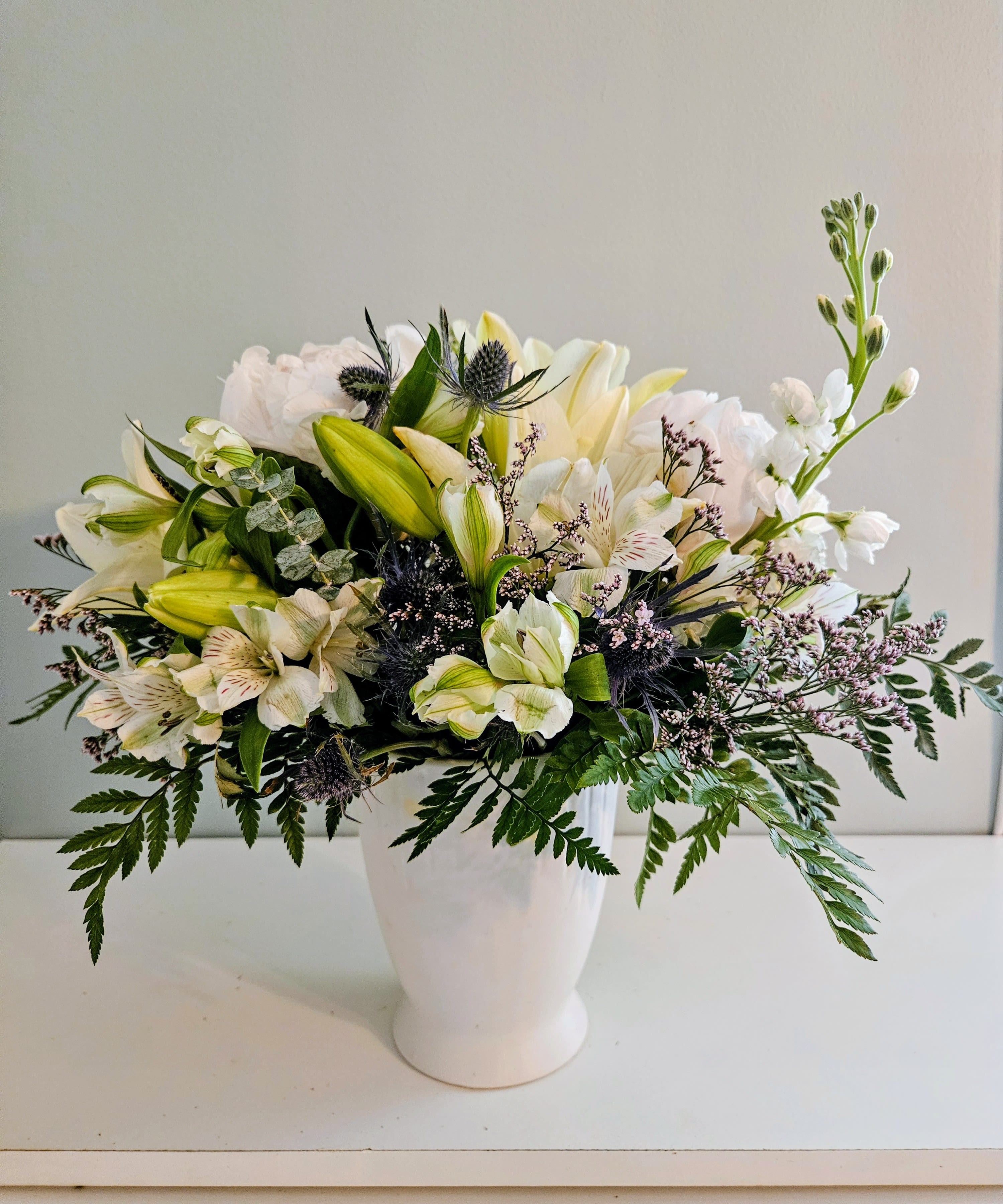 Day Of Consolation - Soft white lilies in bud and bloom give this sympathy-style flower arrangement a quietly uplifting presence-perfect as a Day of Consolation bouquet for a funeral, to offer comfort, or as a gentle get-well gift. The rounded, garden-style mound reads calm and elegant, sending a message of soothing support to the person you're thinking of.  Crafted in a white ceramic vase, the design pairs large white lilies with pale yellow and white alstroemeria, small filler blossoms, airy fern-like foliage, and darker thistle-like accents for subtle contrast and texture. The palette of white, green, yellow, and a touch of pink creates a fresh, understated look; the low, full shape makes it ideal for tabletops or a memorial display.  This arrangement feels like a heartfelt way to say you care-suitable for funerals, bedside visits, or to comfort someone at home. The compact vase keeps water contained and display-ready; for best longevity, keep blooms in a cool spot away from direct sun and refresh the water every couple of days.