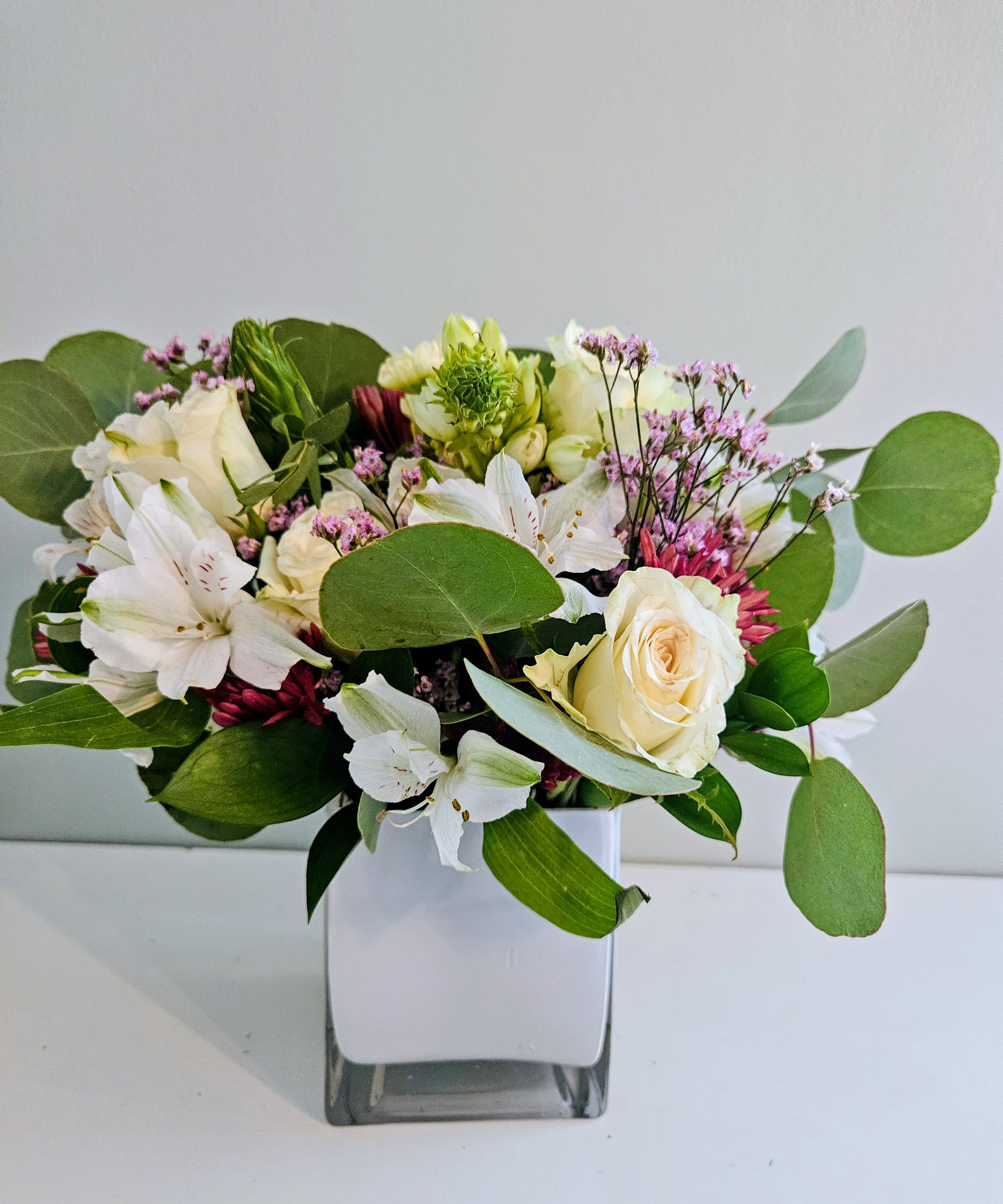 Peace Of Calm - Soft cream roses and white alstroemeria mingle with delicate pink filler blooms to create a Peace Of Calm flower arrangement perfect for birthdays, thank you gifts, or a gentle thinking-of-you gesture. The calming palette of white, green, and pale pink gives the bouquet an elegant, peaceful presence that will help the person you're sending it to feel seen and soothed.  Set in a compact square glass vase, the design is framed by large, round eucalyptus-like foliage that adds an airy, garden-fresh texture around the blooms. The arrangement is intentionally compact yet full, with cream roses as the focal points, clusters of white alstroemeria for softness, and small pink accents for a whisper of warmth-an understated look that complements both home and office settings.  This everyday arrangement arrives ready to display in its clear glass vase; it's the kind of gift that quietly says you care. Ideal for someone who prefers a refined, natural aesthetic, it makes a calming centerpiece or bedside bouquet and is easy to keep fresh with regular water changes and trimming the stems every few days.