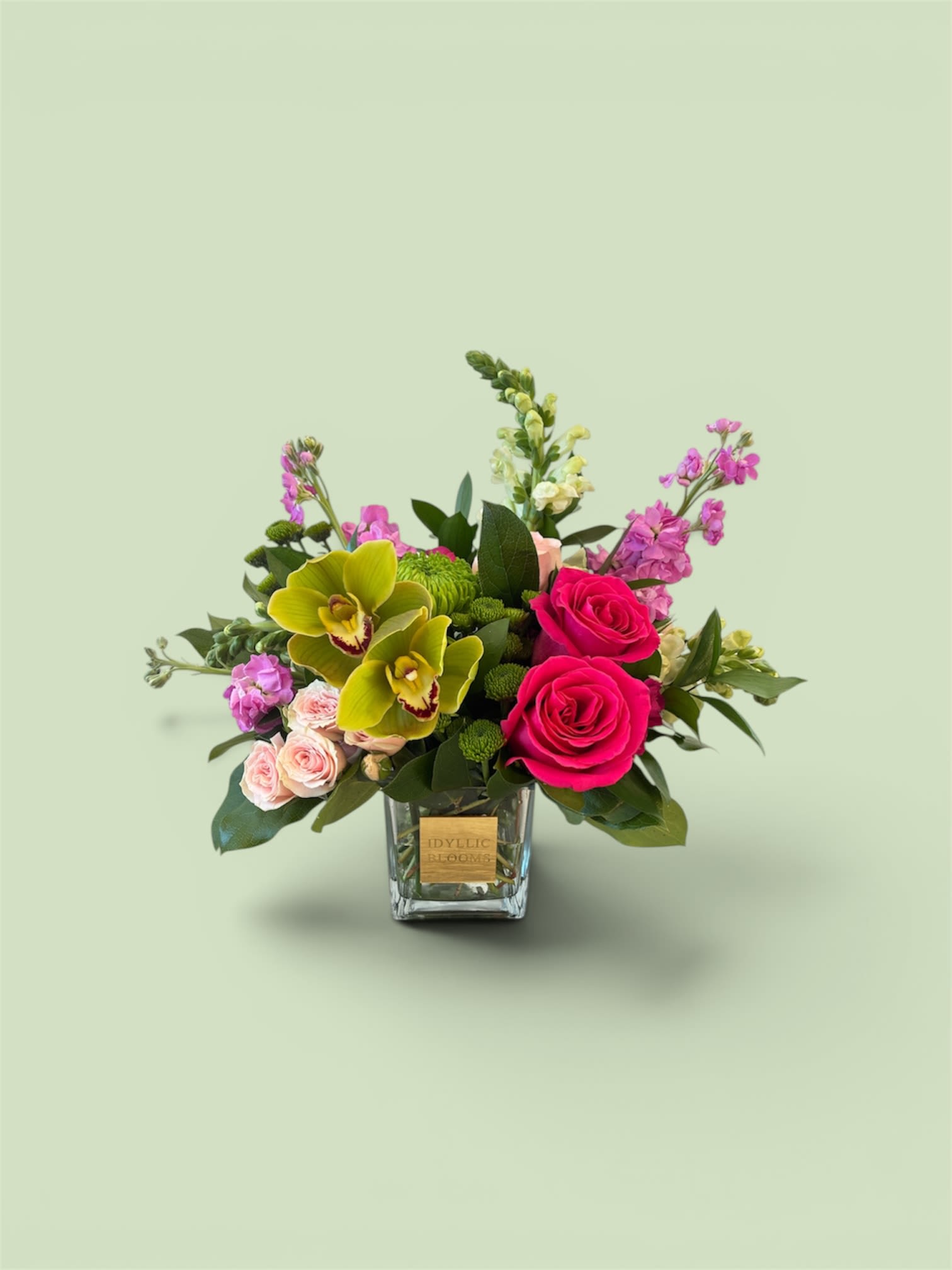 Vibrant garden LUXE - Vibrant Garden Luxe is a bold and joyful arrangement bursting with color and texture. Featuring striking hot pink roses, exotic green cymbidium orchids, soft blush spray roses, and playful snaps and stock, this design brings together elegance and energy in perfect harmony. Lush greenery and modern styling in a glass cube complete the look, making it a stunning statement piece for celebrations, birthdays, or anyone who loves a pop of color.