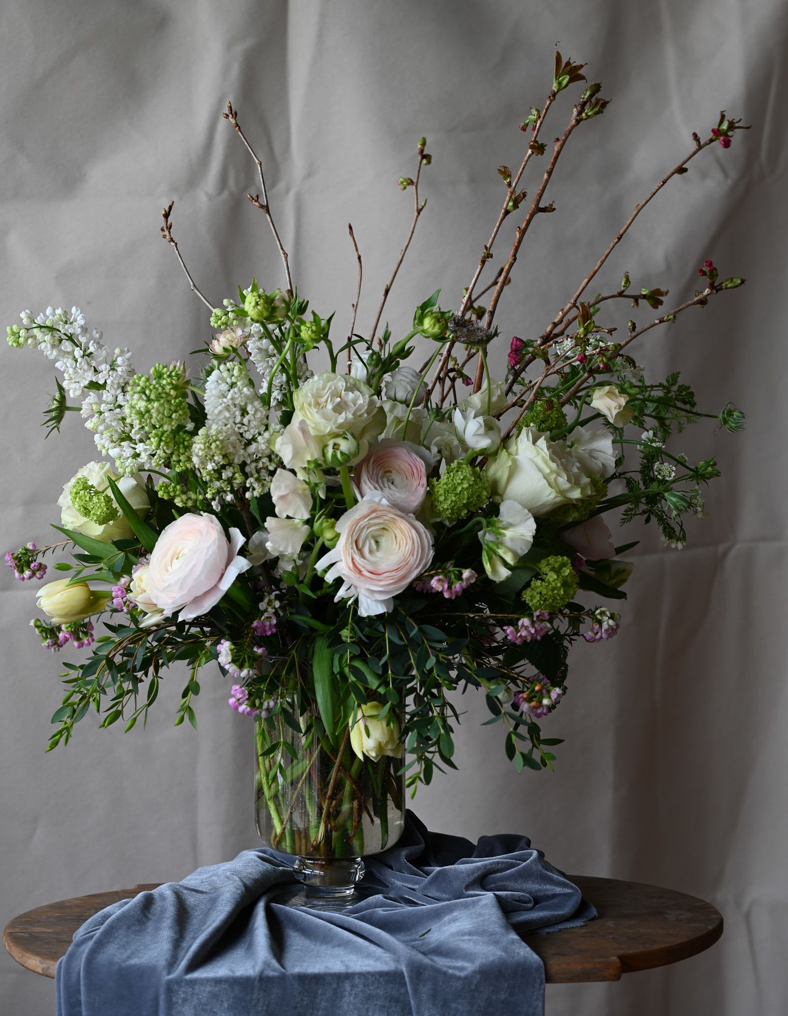 Springtime in the Garden - Flowering branches of lilac, cherry blossom, premium ranunculus, garden roses as well as other seasonal garden flowers enhance this beautiful composition of spring in a footed hurricane vase.  
