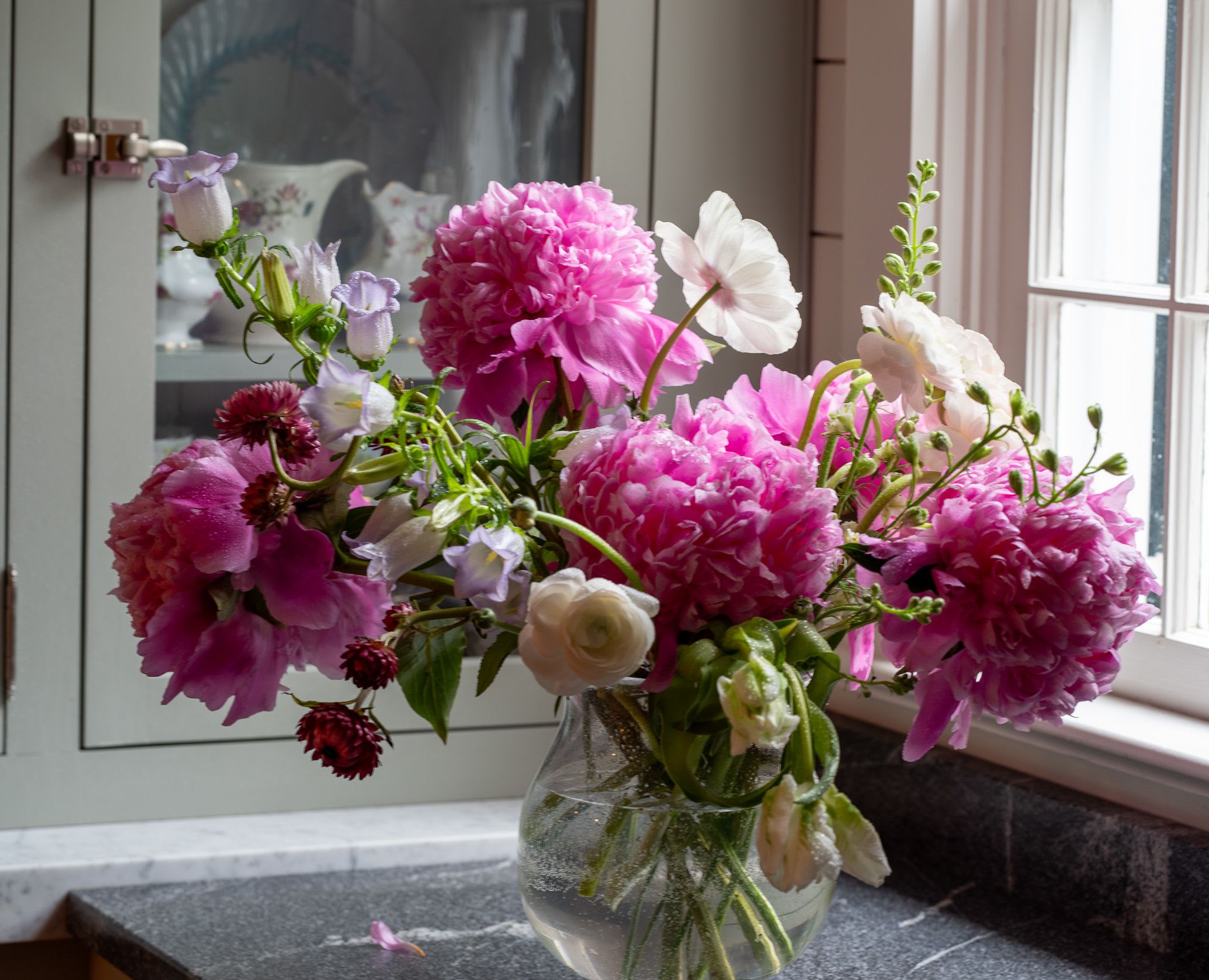 Loose arrangement of pink peonies and white blooms in a clear glass vase on a countertop.