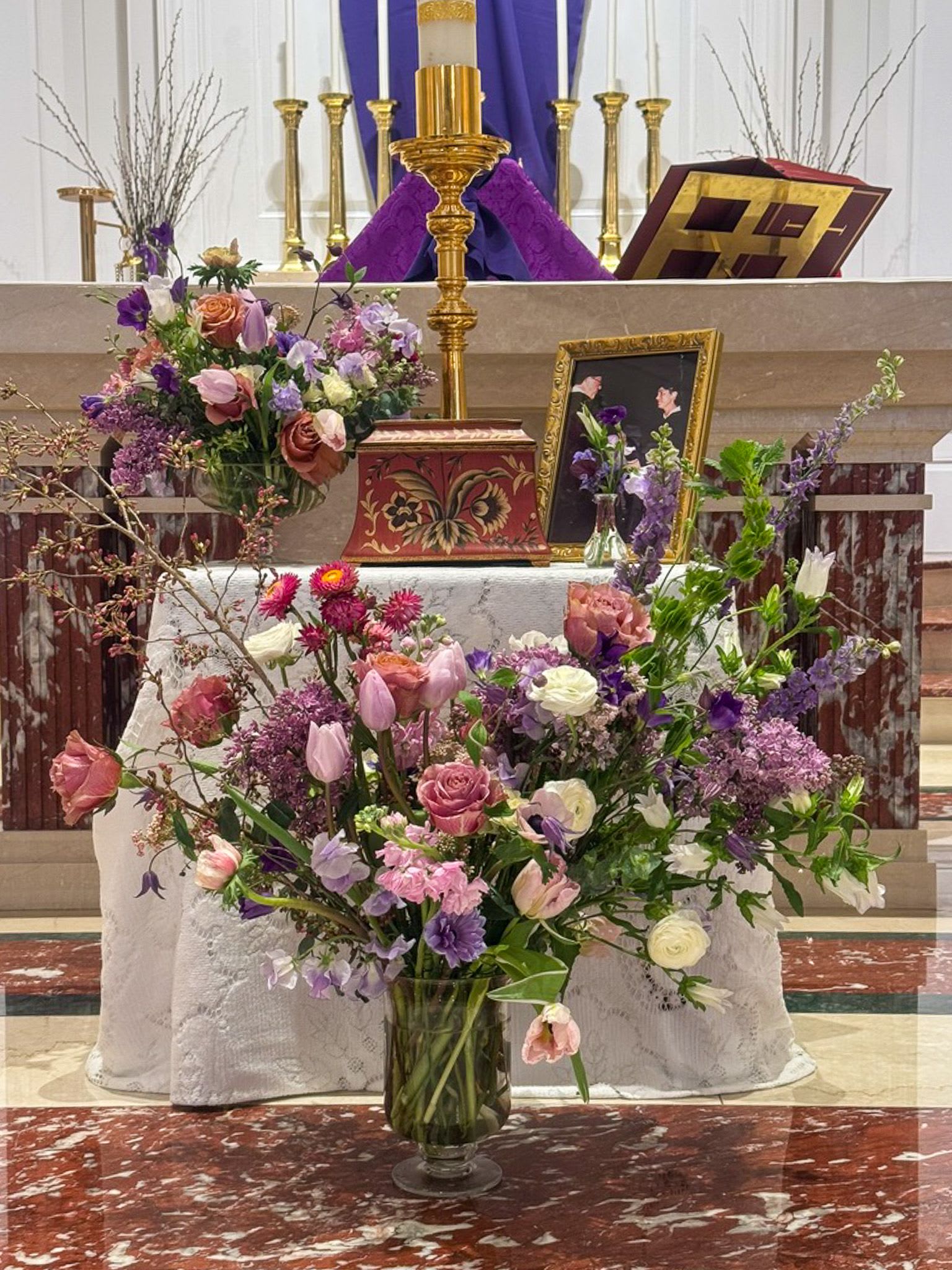Large purple and pink mixed flower arrangements on a church altar with candles and framed photo