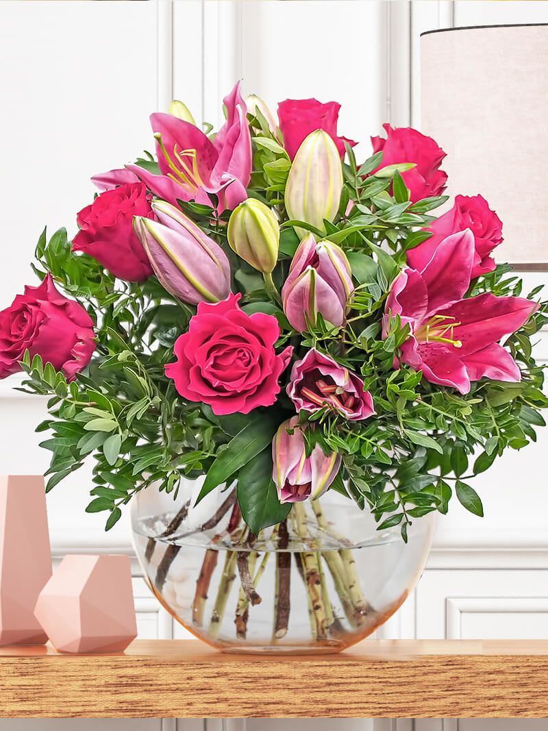 Arrangement of hot pink roses and pink lilies in a round clear glass vase on a wooden shelf