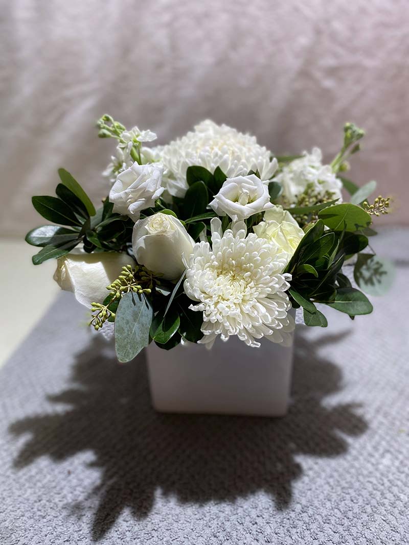 White floral arrangement in a square white vase with dark green foliage
